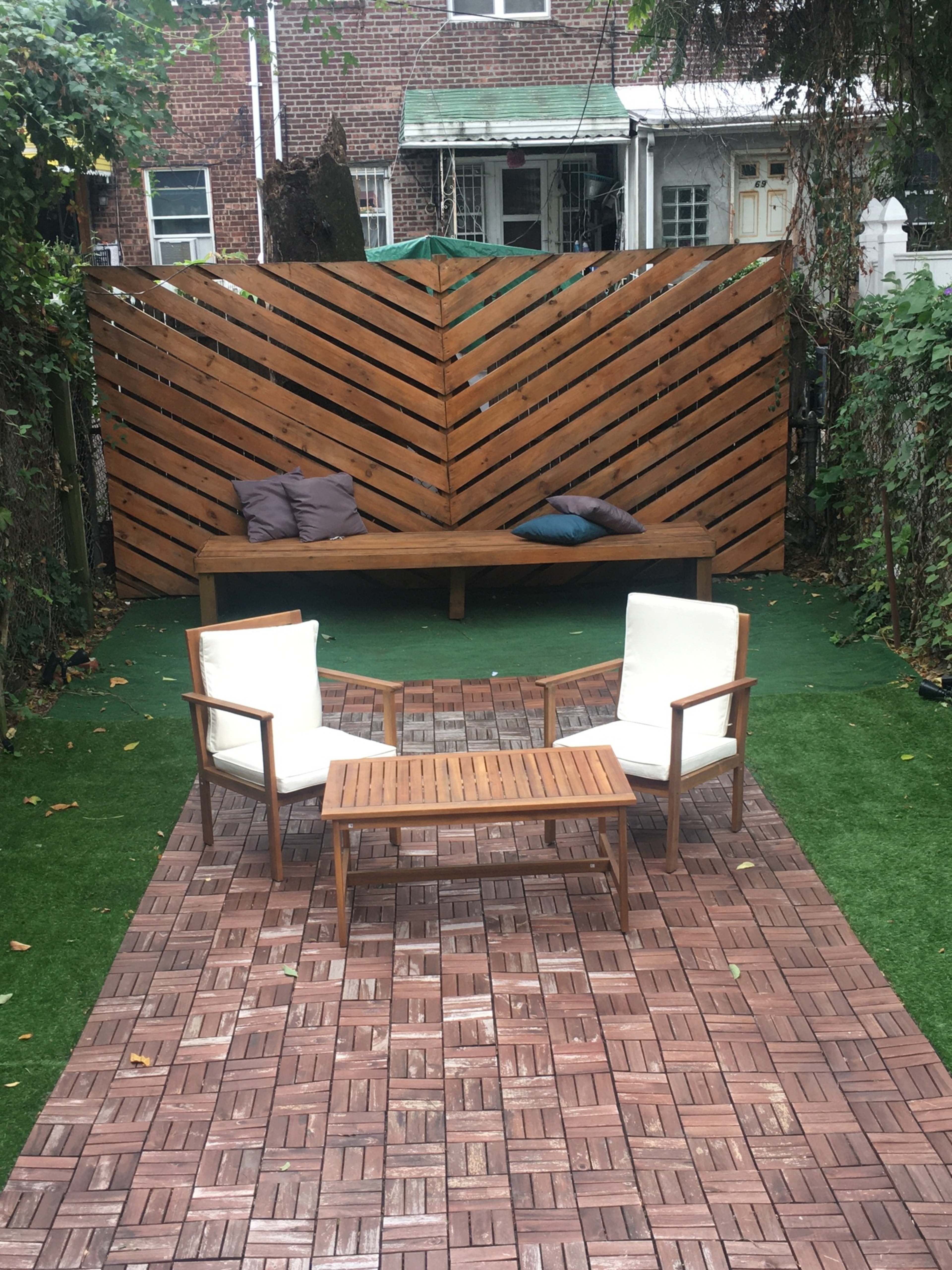 A backyard seating area featuring two white armchairs and a wooden coffee table on a tiled surface, backed by a slatted wooden wall and surrounded by greenery.