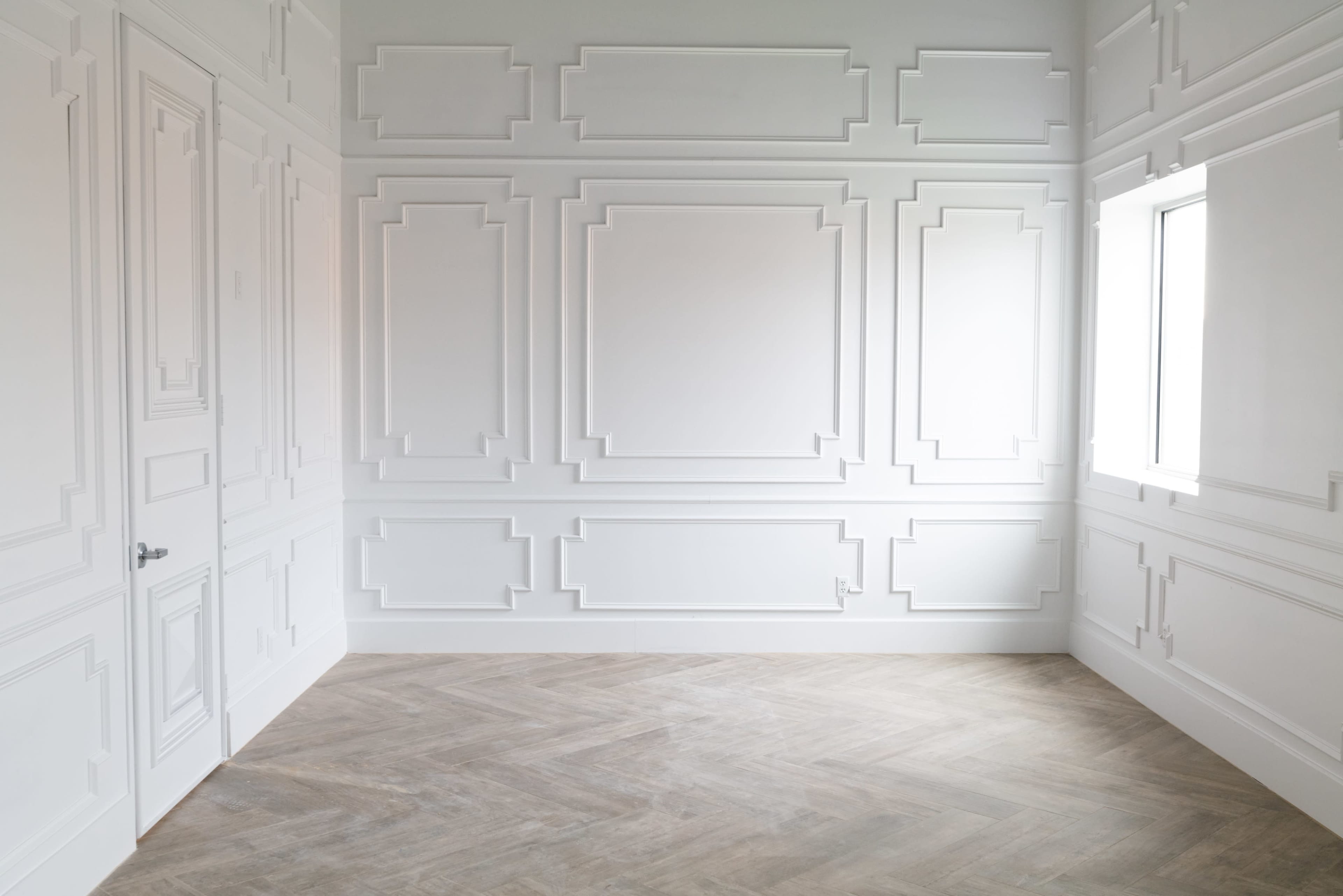 A minimalist room with white paneled walls and a window, featuring wooden herringbone flooring.