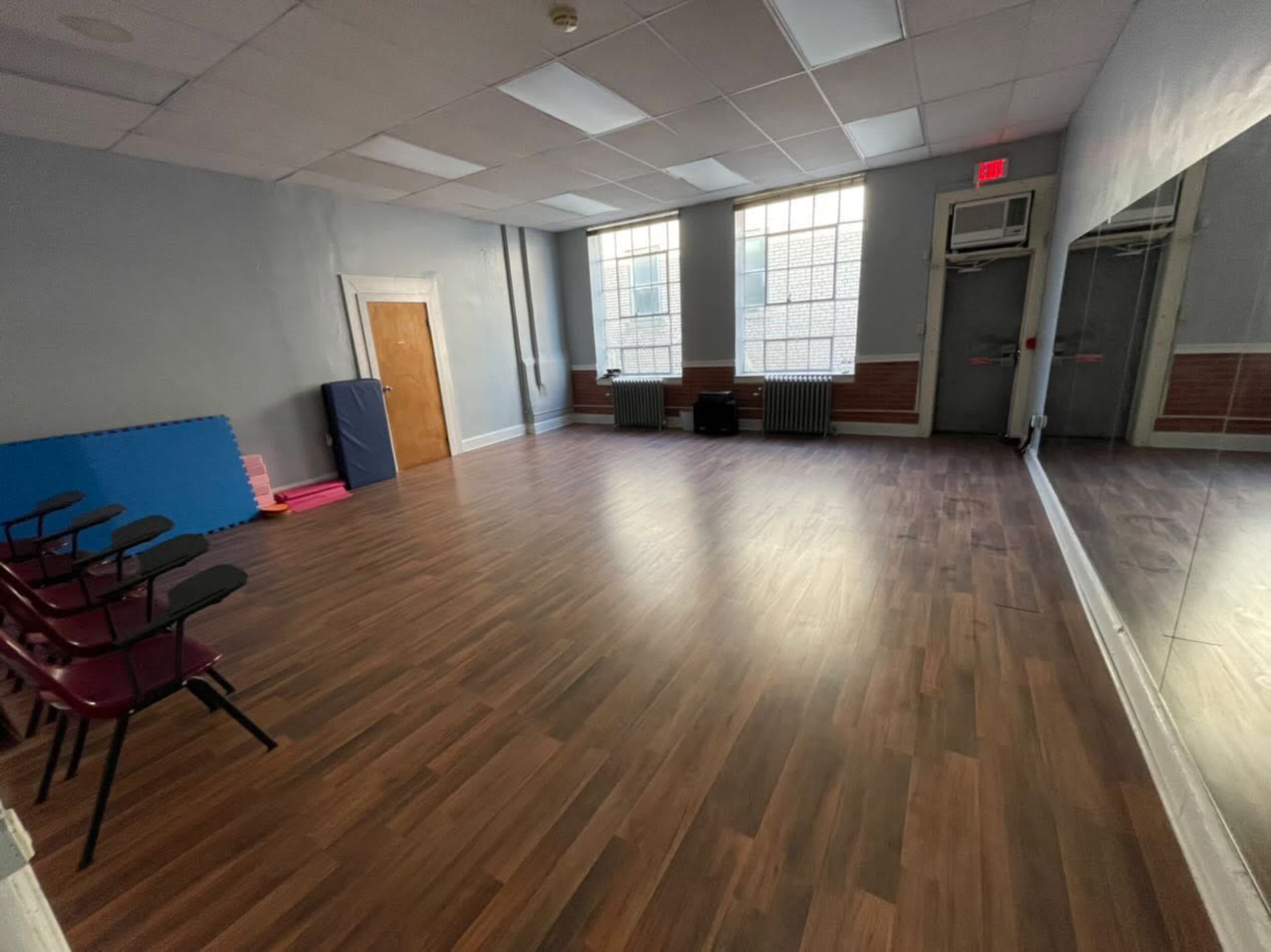 A vacant room with wooden flooring, large windows, and a mirrored wall, furnished with a few stacked chairs and exercise mats.