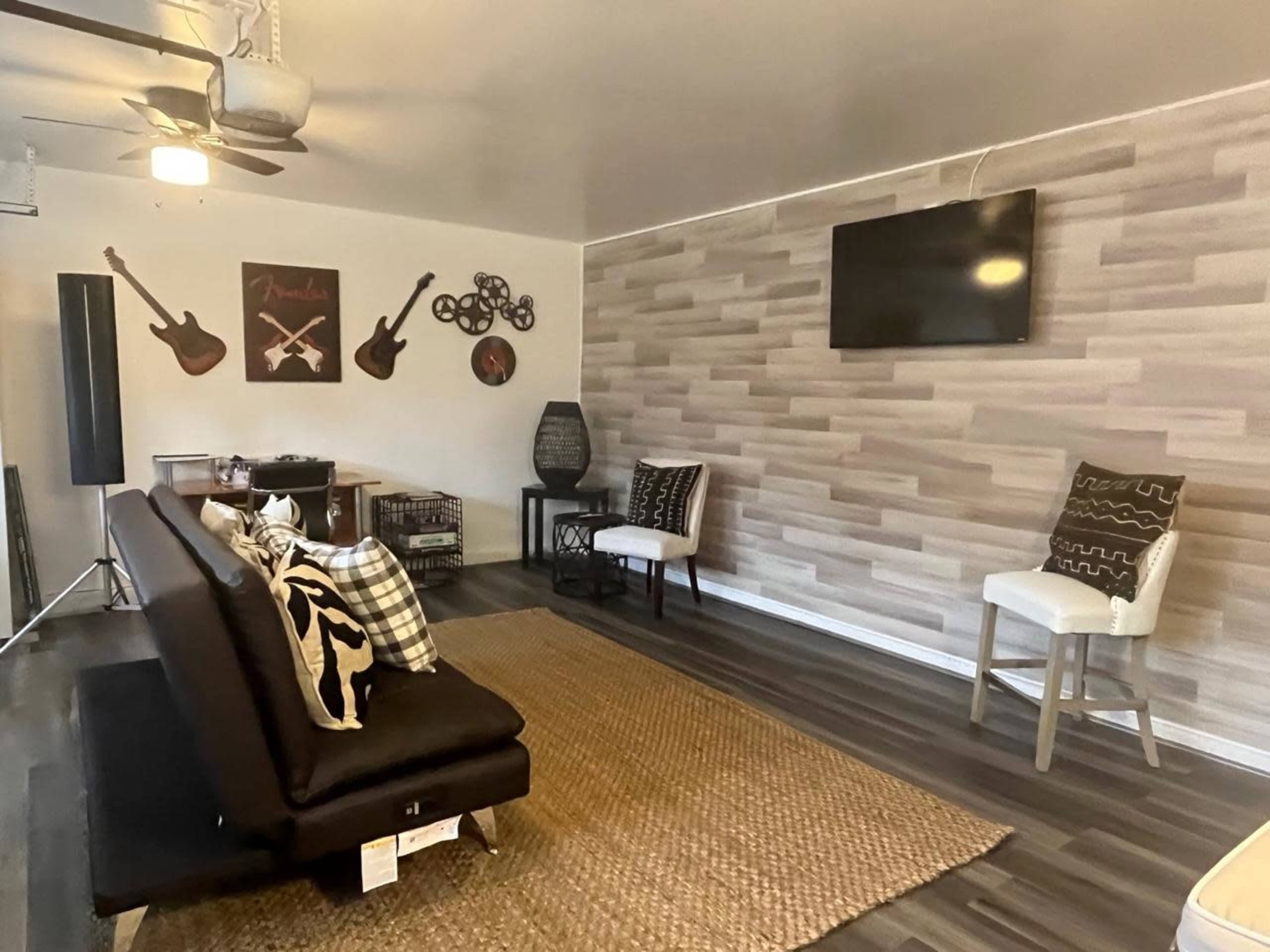 A living room with a black futon sofa, a woven rug, a wall-mounted television, and decorative items including guitars and a modern light fixture.