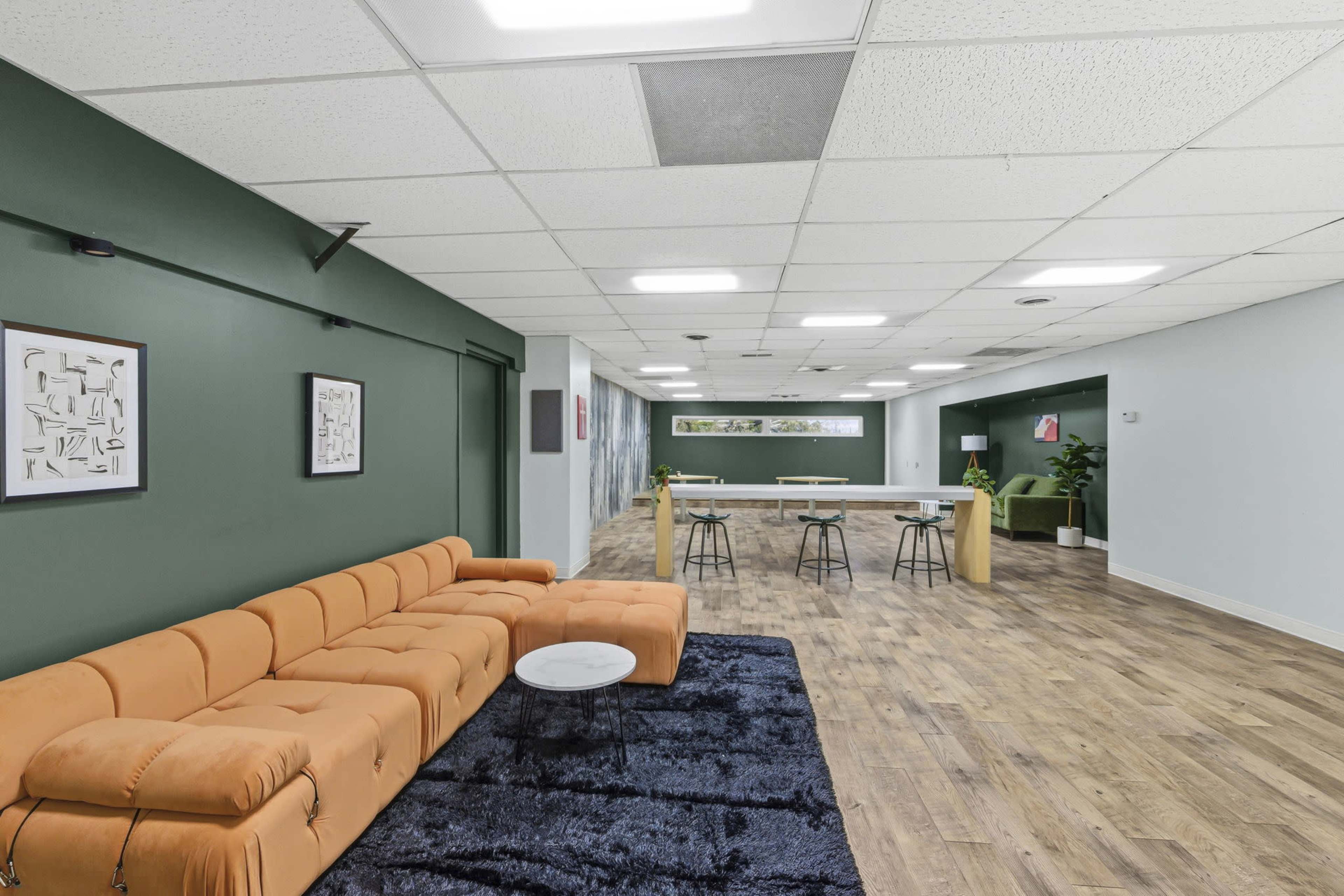 The image shows a modern, open shared workspace featuring a large orange couch, a black rug, and a bar-style table area with chairs against a green and grey wall.