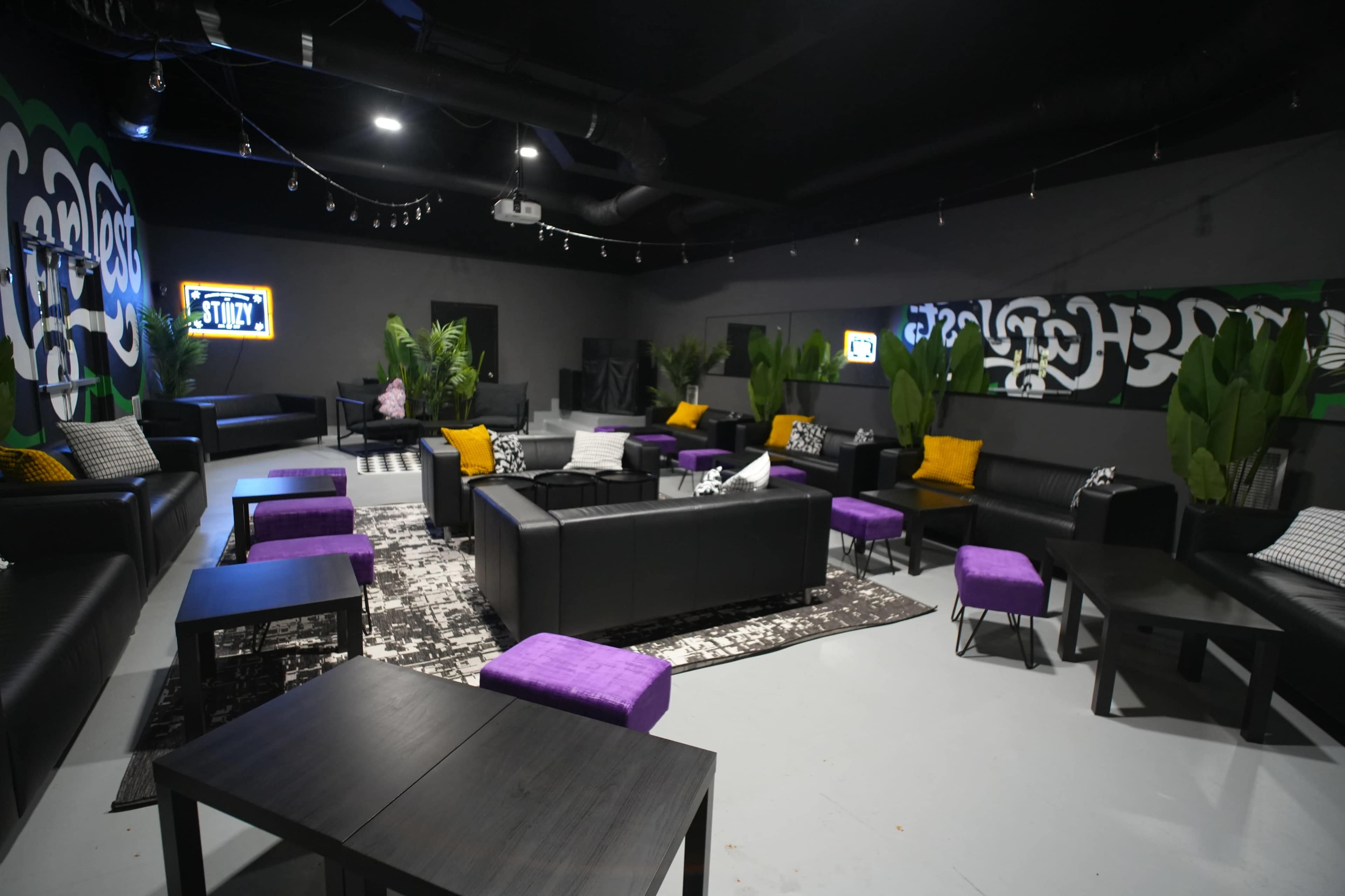 The image shows a modern lounge area with black sofas, purple ottomans, and potted plants, arranged around low tables in a spacious room with colorful wall art.