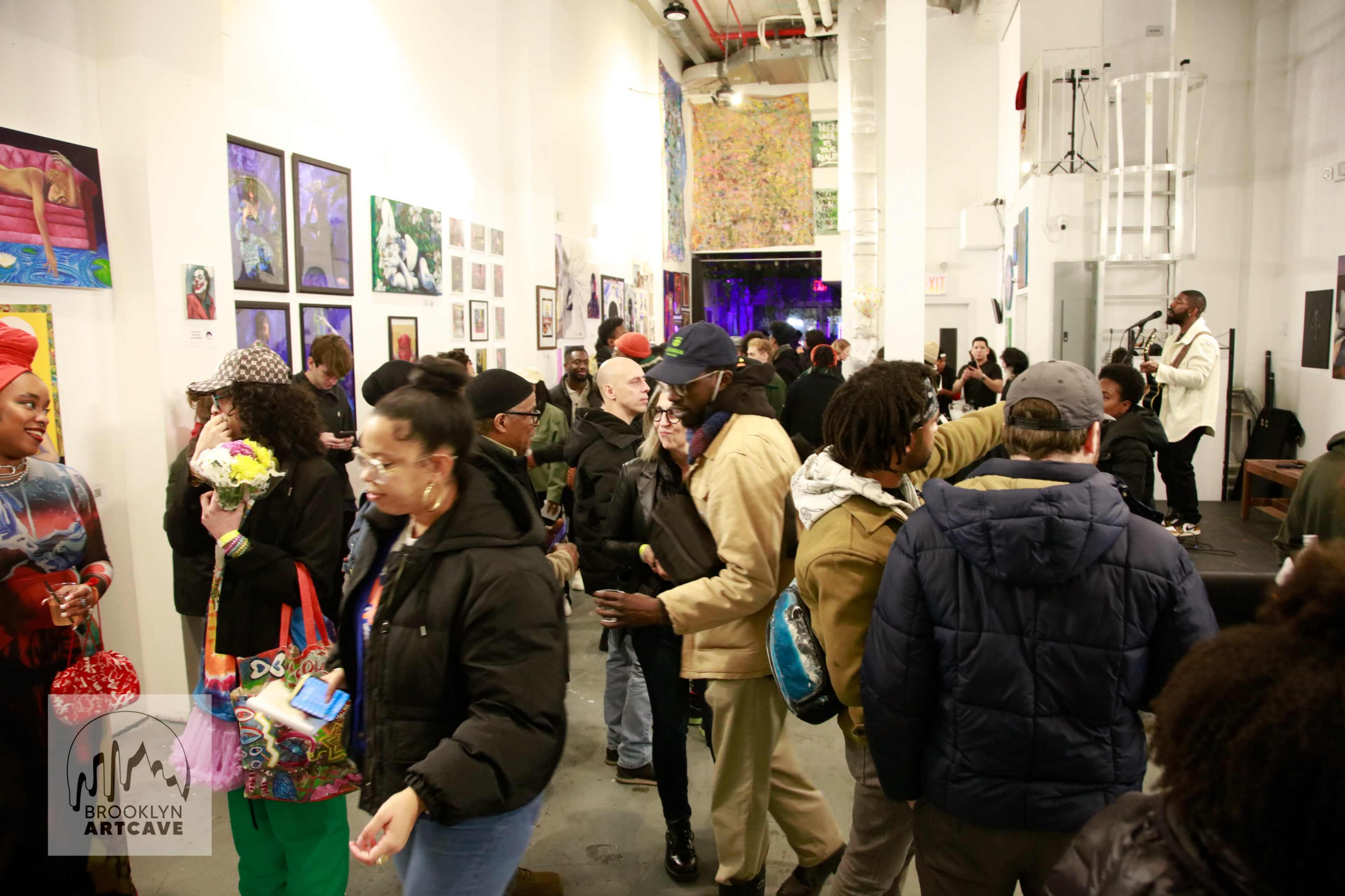 A crowded art gallery features various artworks on the walls and attendees mingling, with a performer on a small stage in the background.
