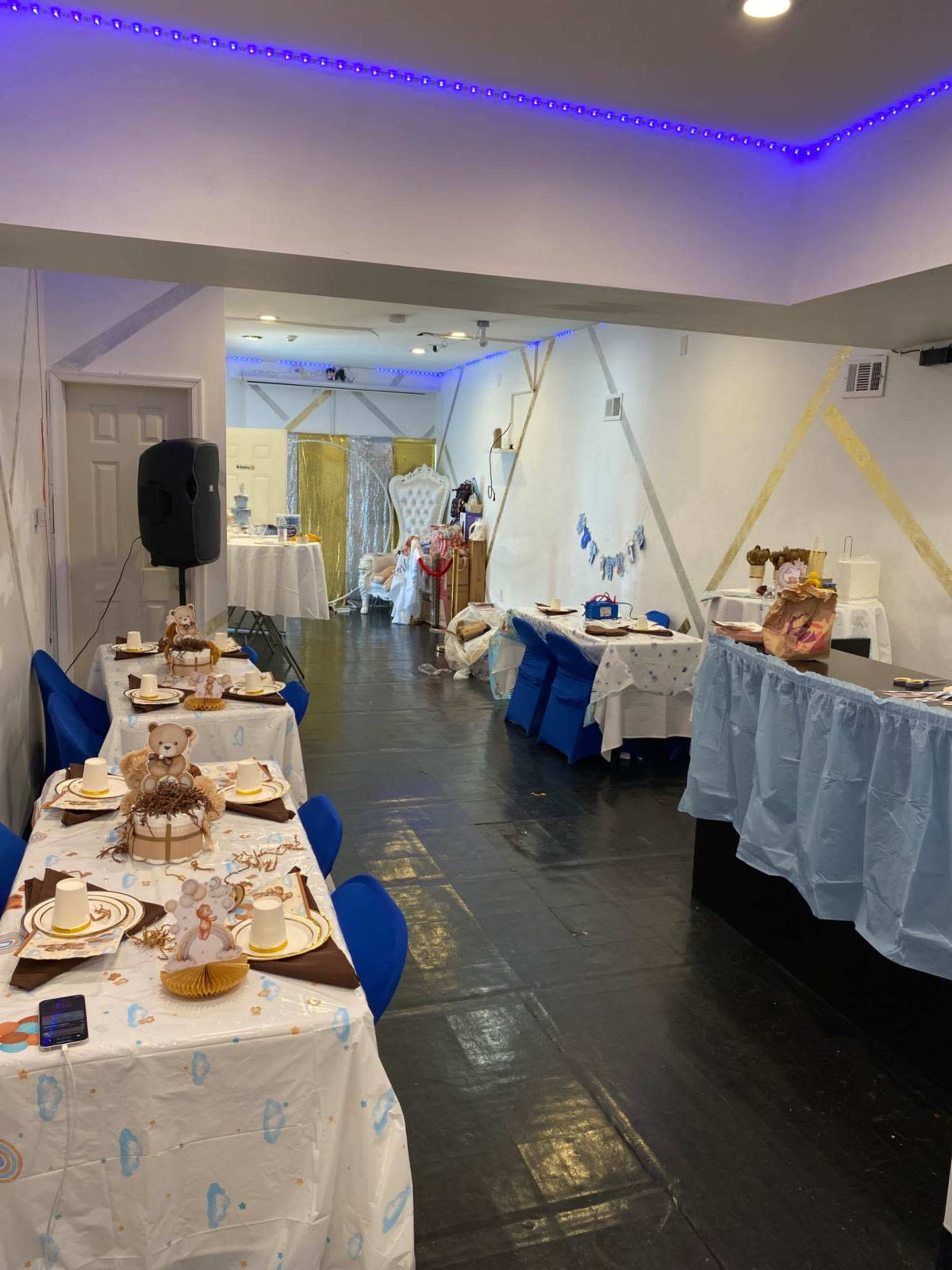 The image shows a decorated indoor party space with multiple tables set for guests, each featuring tableware, and a backdrop of streamers and decorations along the walls.