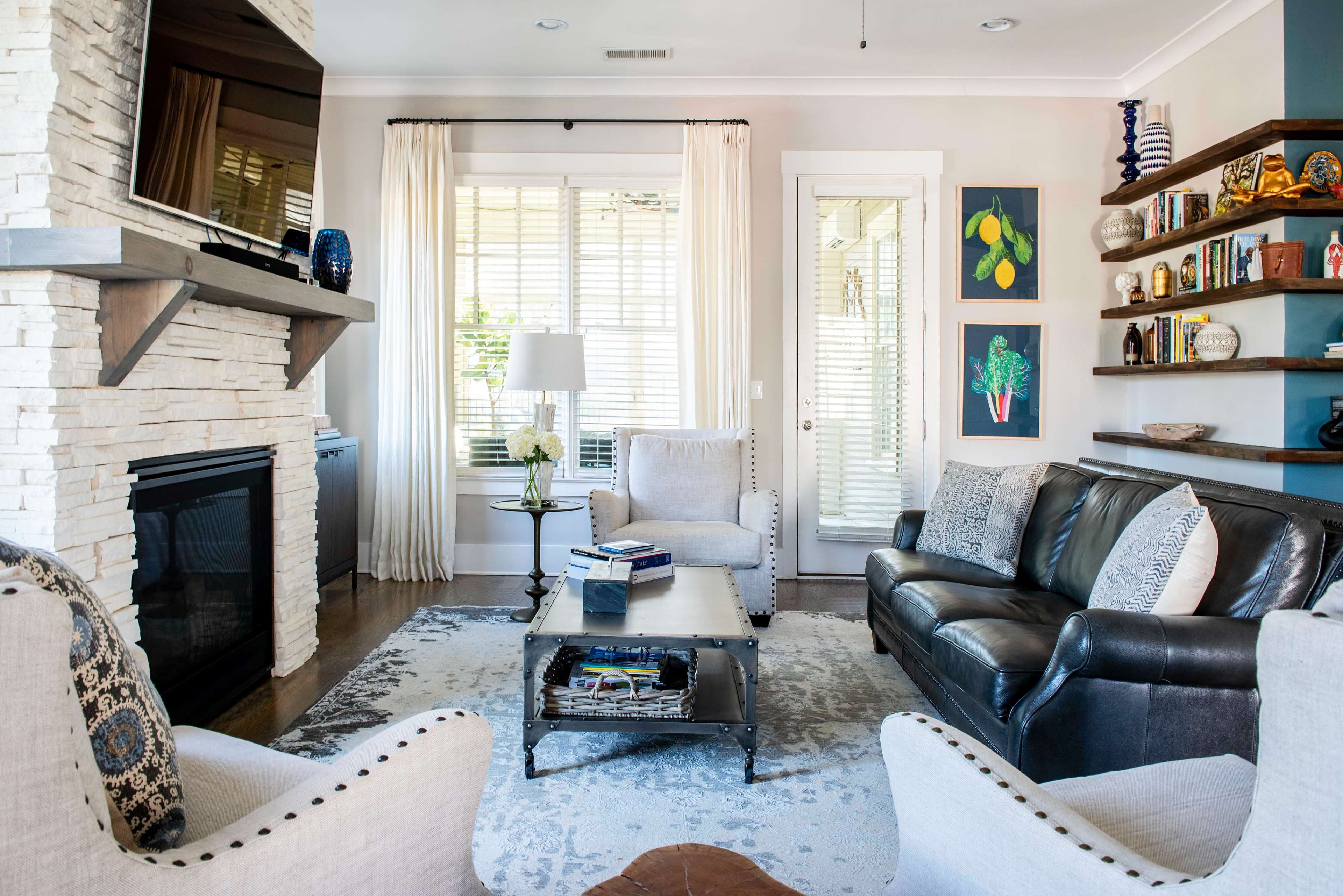 The image shows a modern living room featuring a fireplace with a flat-screen TV above it, two armchairs, a leather sofa, and a coffee table in the center, surrounded by decorated shelves and framed artwork on the walls.
