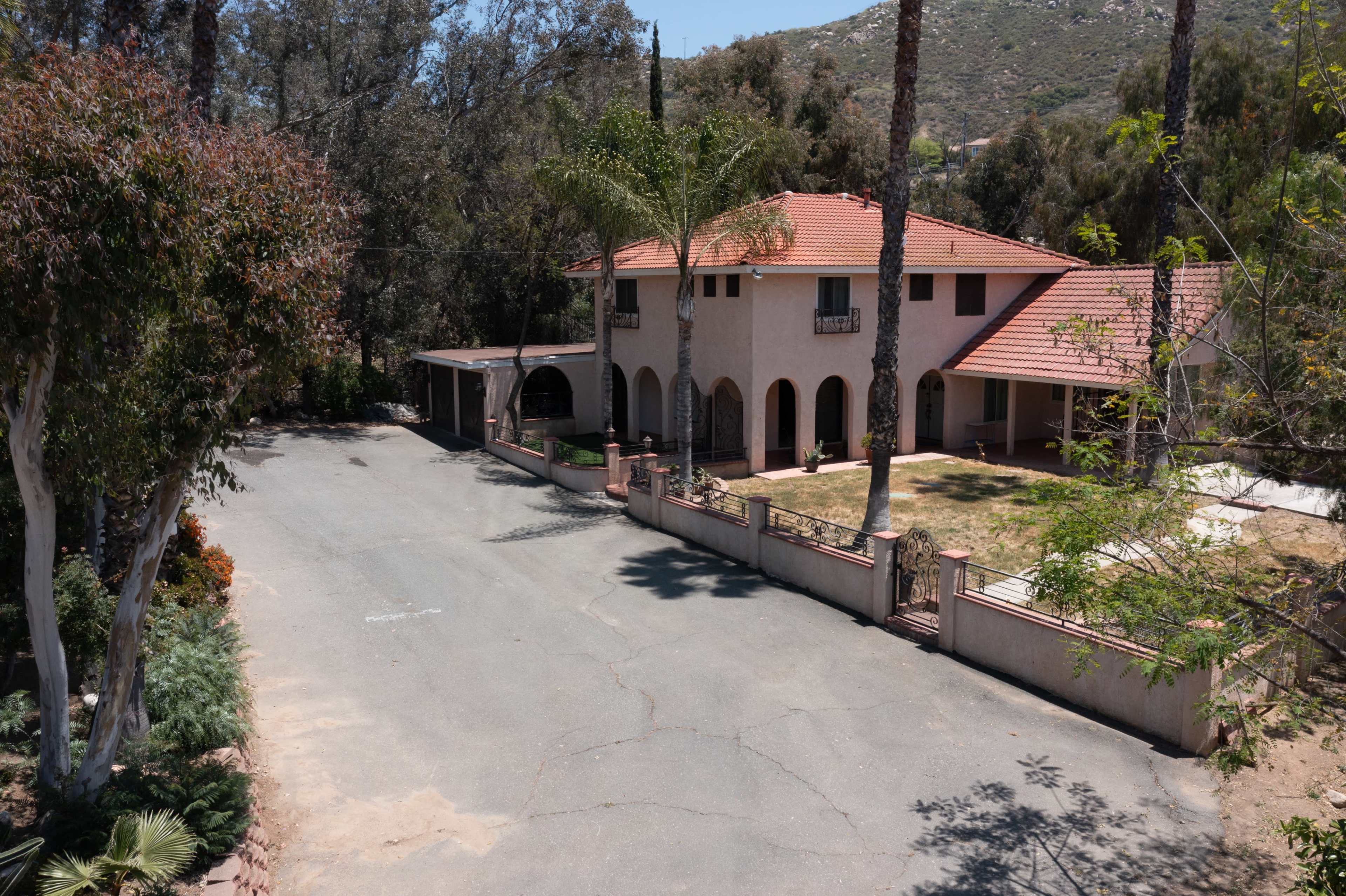 A large two-story house with a red tiled roof is situated at the end of a winding, tree-lined street, with a palm tree and a green lawn in the front yard.