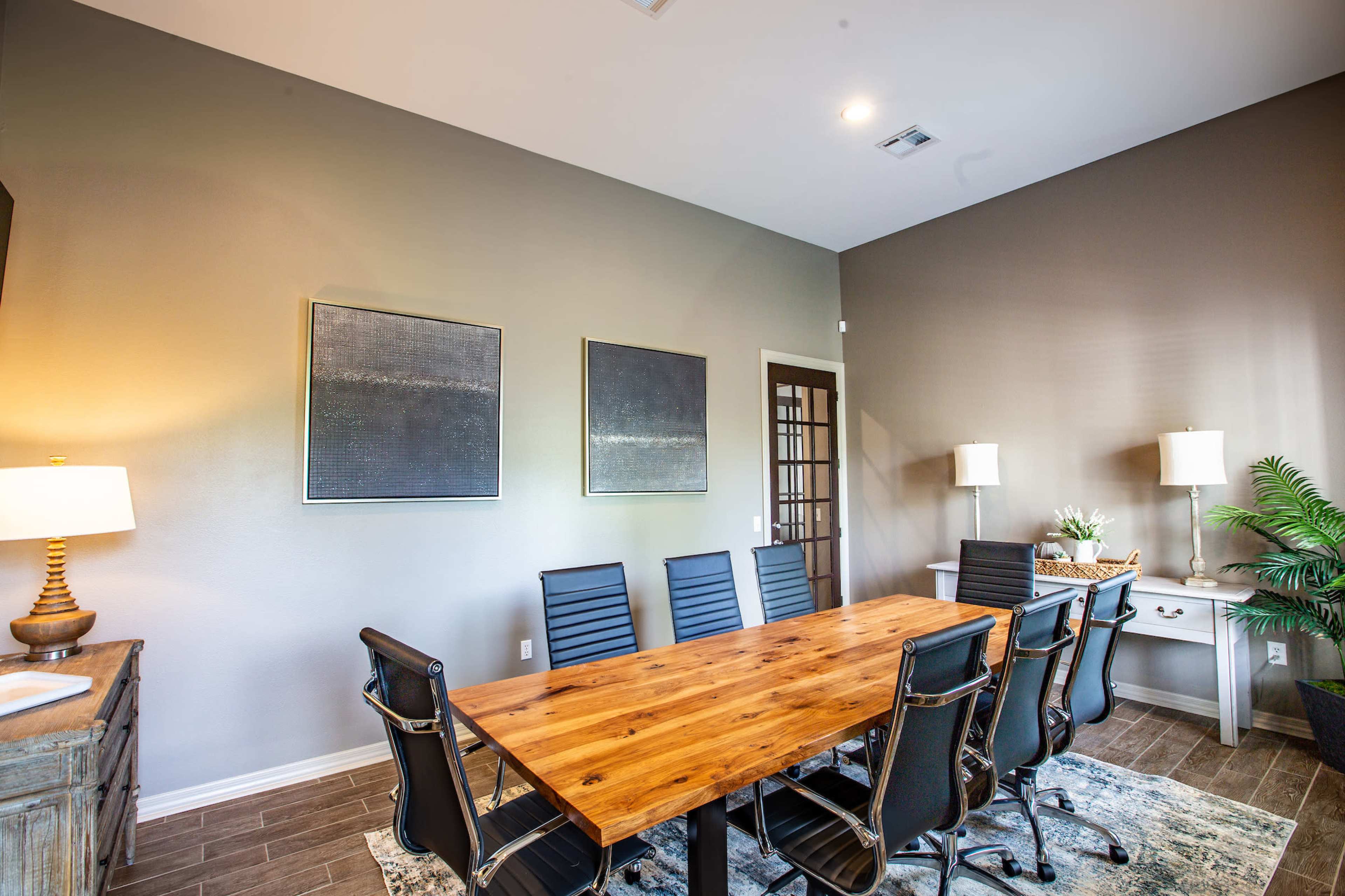 A rectangular wooden conference table with black mesh chairs is set in a meeting room featuring framed artworks on the wall and a side console with decorative items.