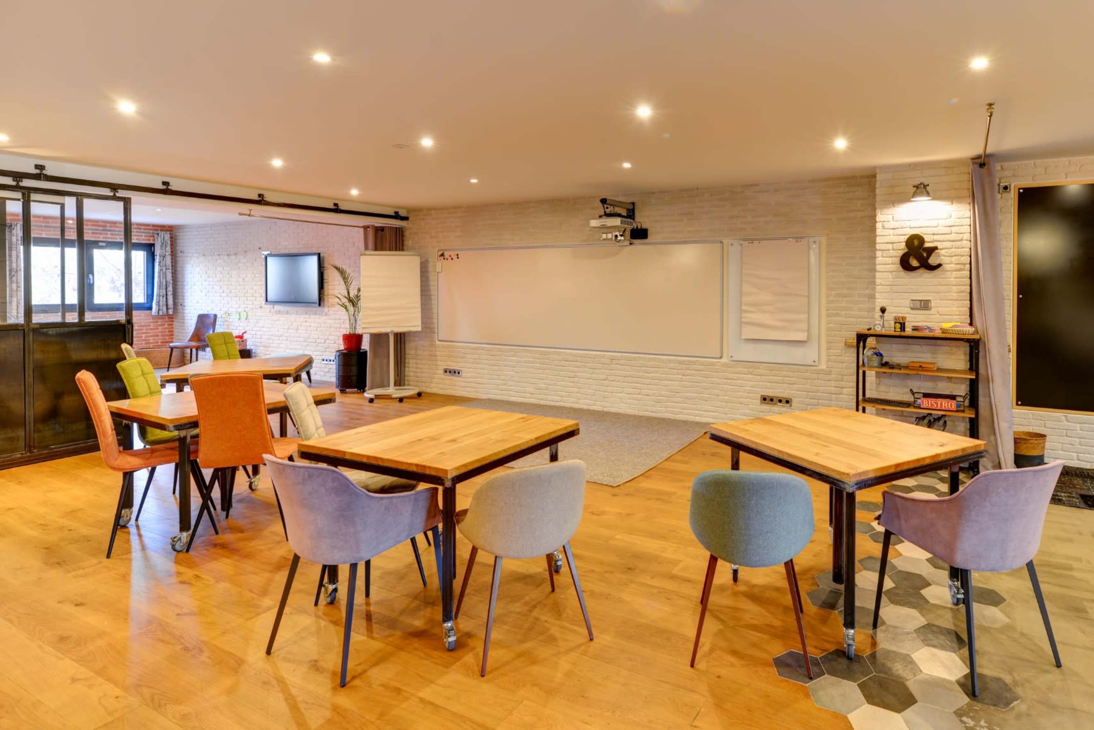 A modern classroom features multiple tables and chairs arranged on a wooden floor, with a whiteboard and projector visible on one wall.