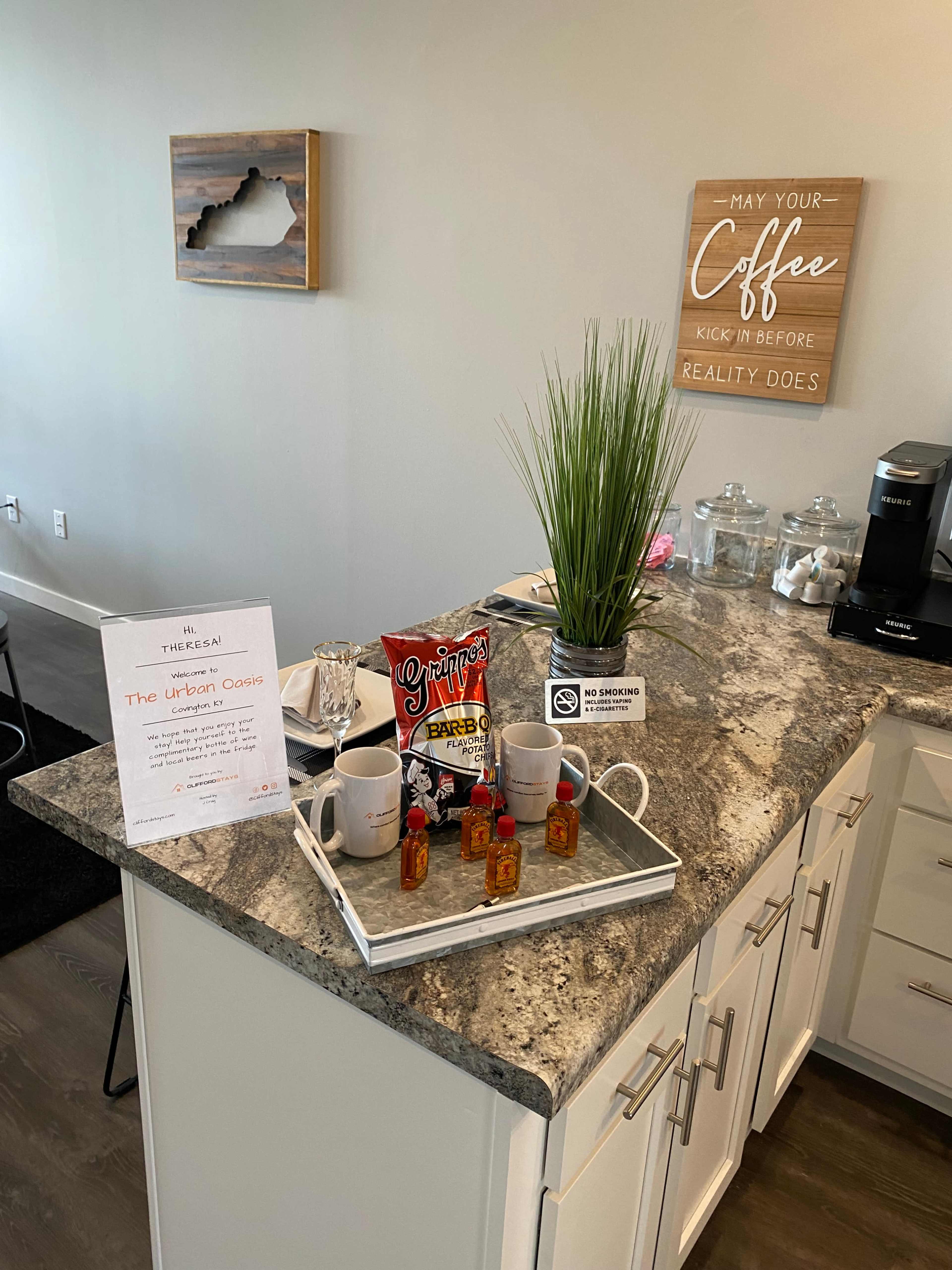 A countertop displays a tray with two mugs, small bottles of liquor, snacks, a glass of water, and a sign for "The Urban Cogie" alongside a decorative plant and a coffee machine.