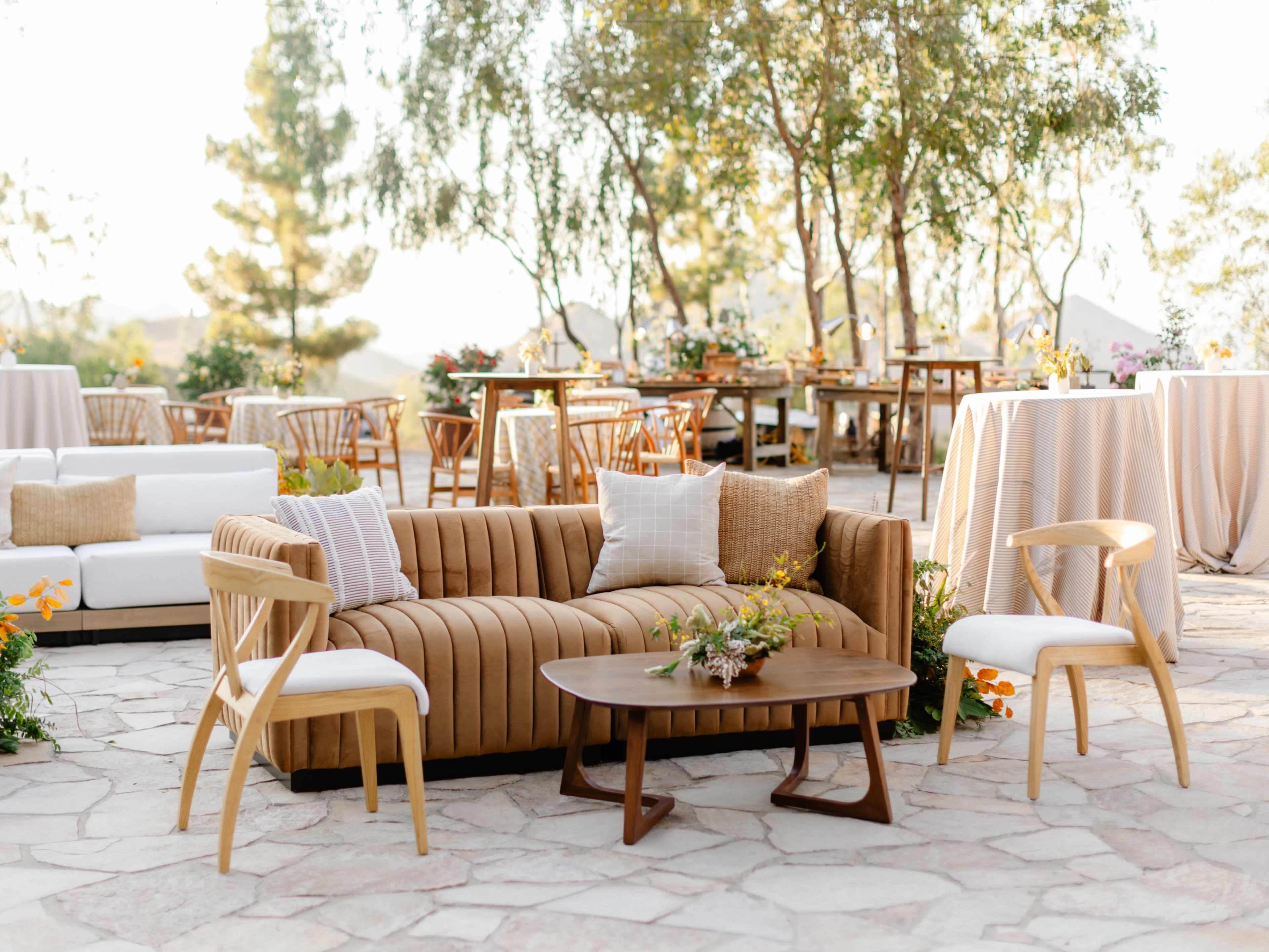 The scene features a stylish outdoor seating area with a brown sofa, wooden chairs, and tables surrounded by trees and floral decorations.