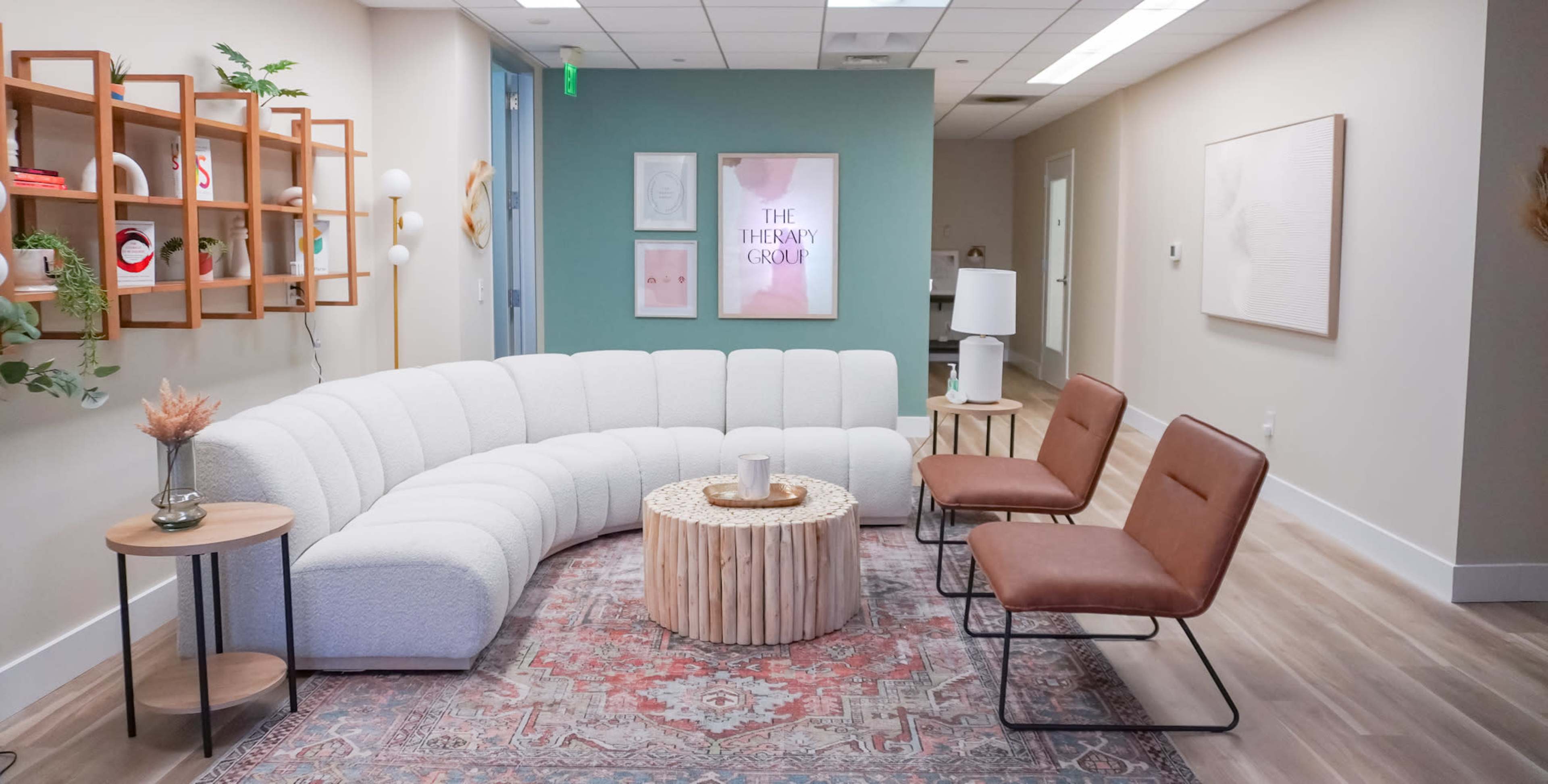 A curved white sofa and two brown chairs surround a wooden coffee table in a well-lit waiting area, featuring light-colored walls and decorative art.