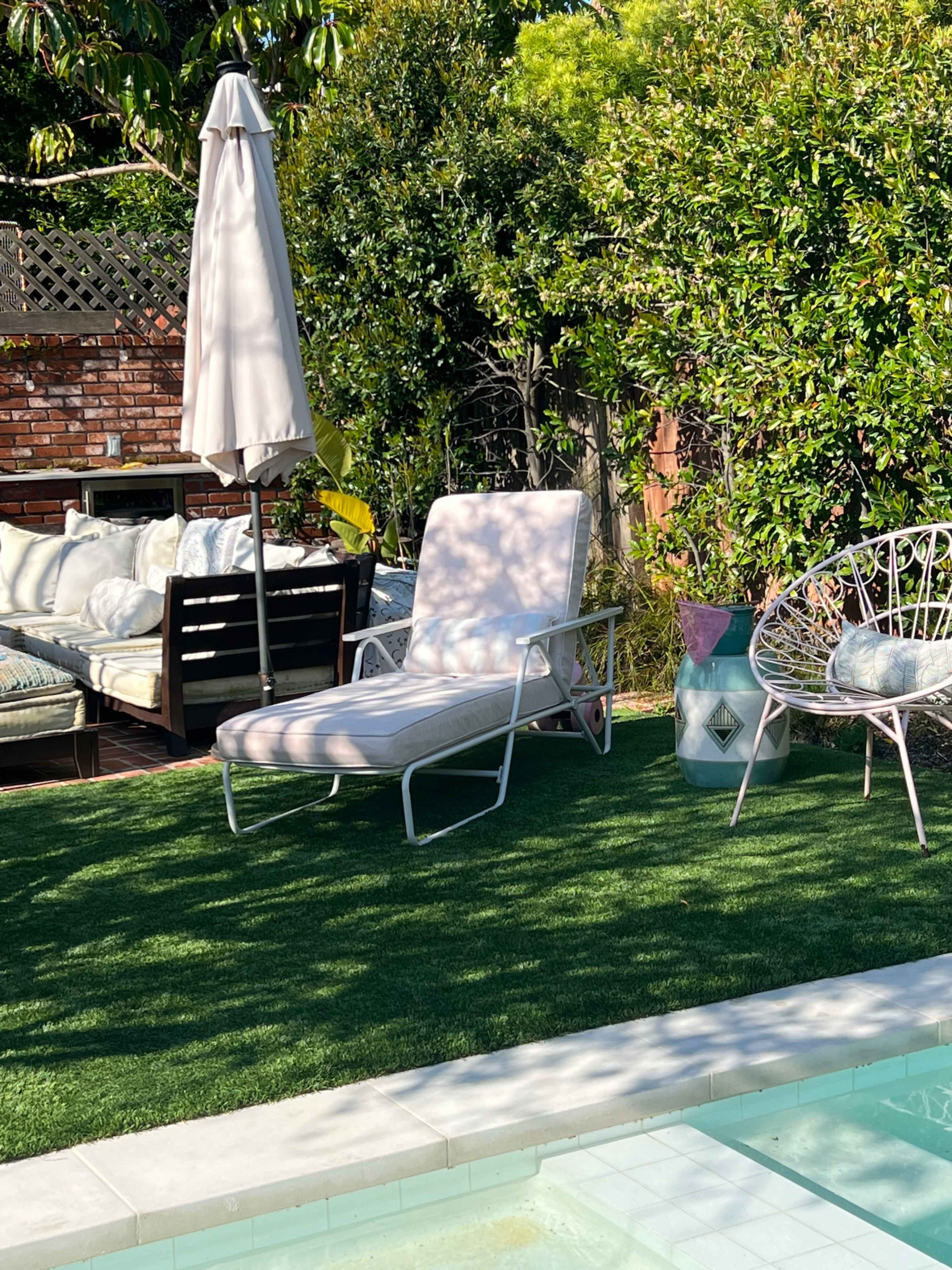 A lounge chair and an umbrella sit on artificial grass beside a swimming pool, with a seating area and greenery in the background.