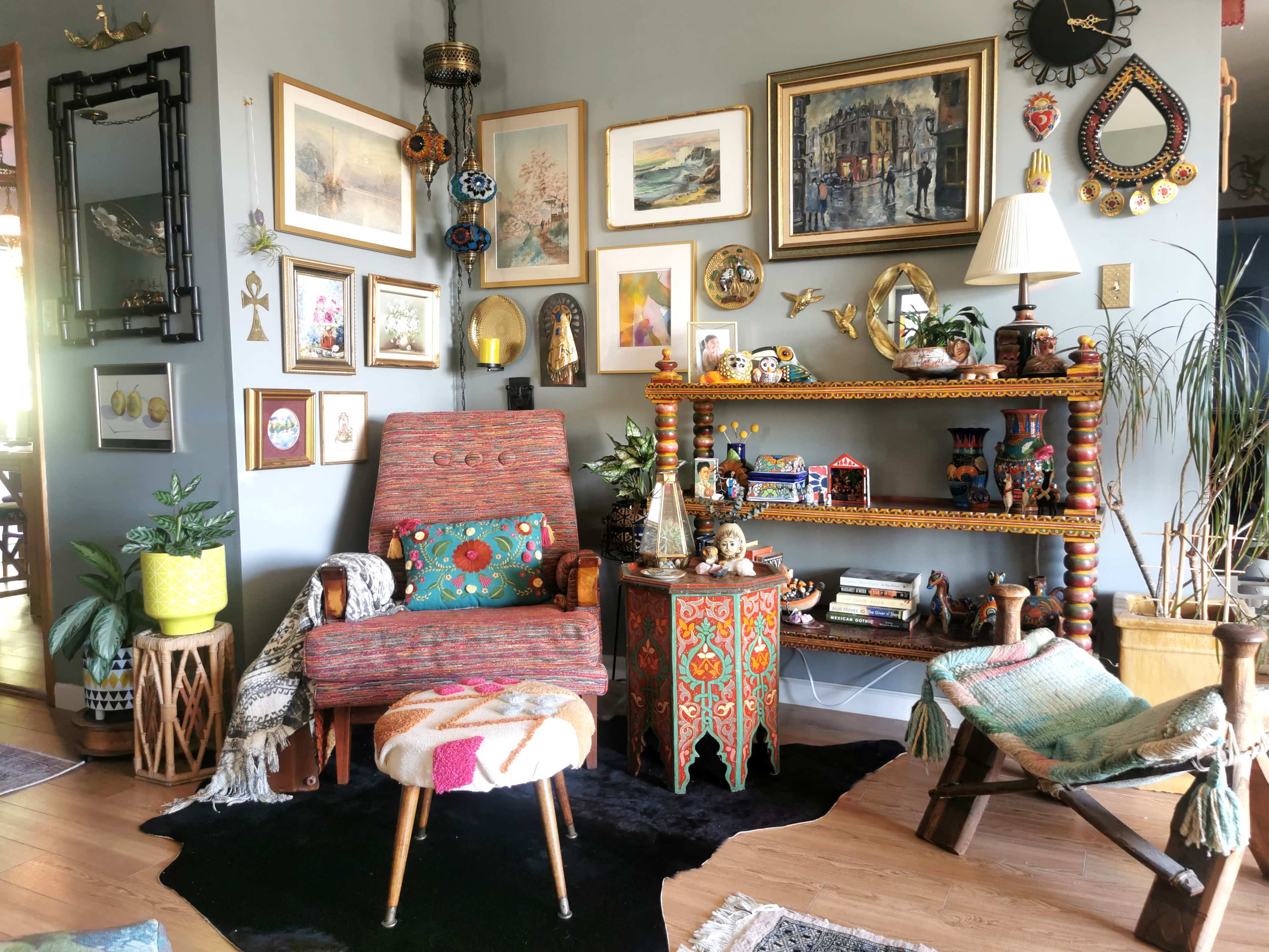The room features a colorful armchair beside a decorative wooden shelf filled with plants, art, and various ornaments, all set against a wall adorned with framed pictures.