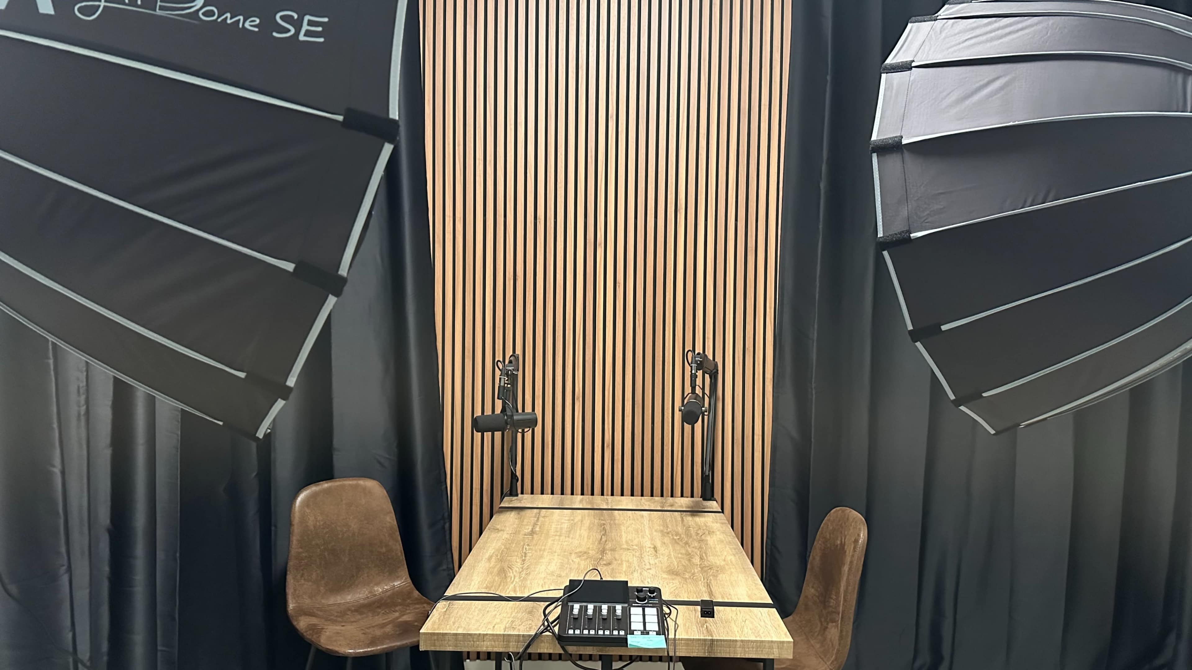 The image shows a podcast recording studio with a wooden table, two microphones on stands, and softbox lights on either side.
