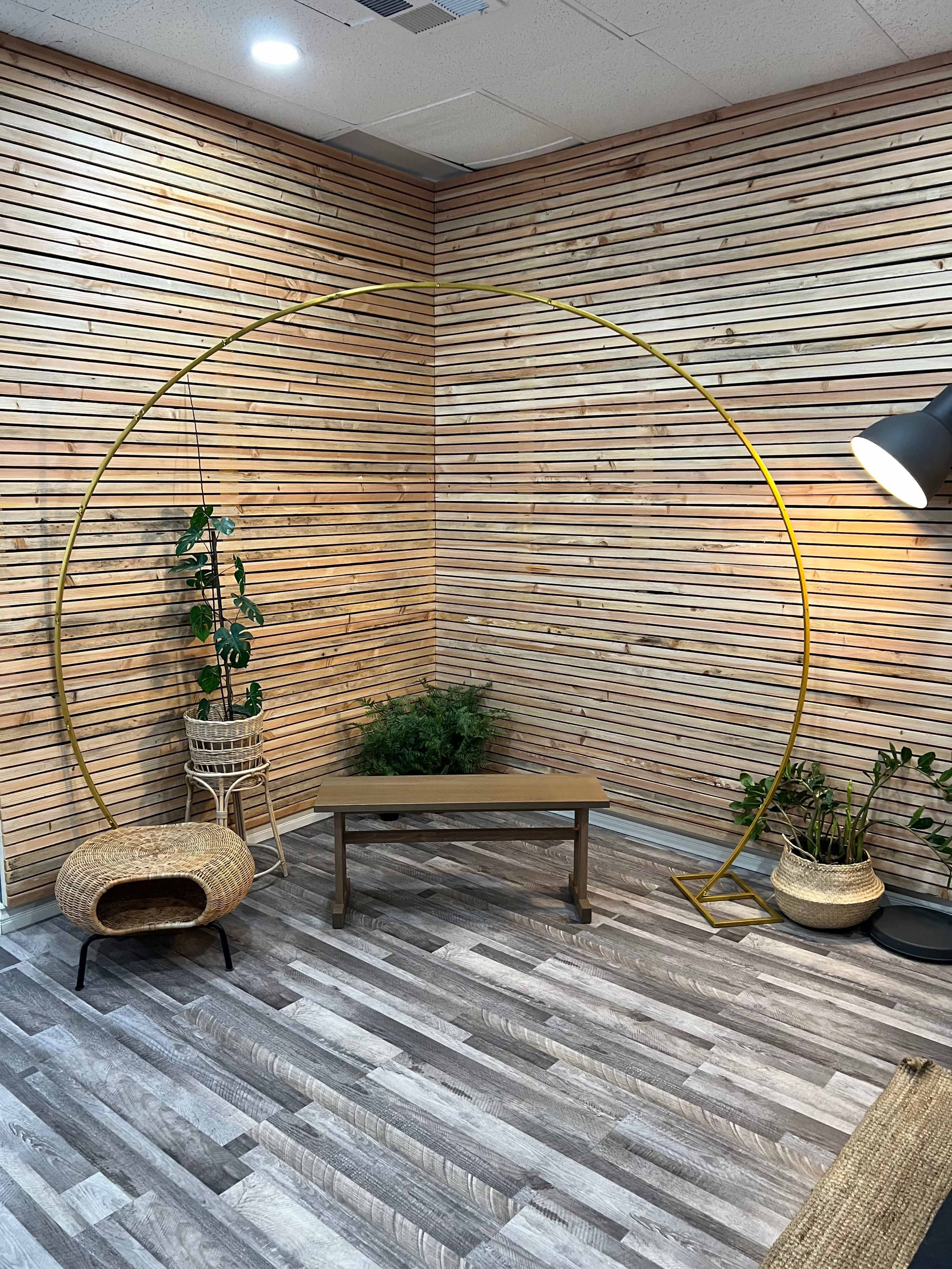 The image shows a corner of a room featuring a wooden slat wall, a yellow circular frame, wooden furniture, and several plants in decorative pots.
