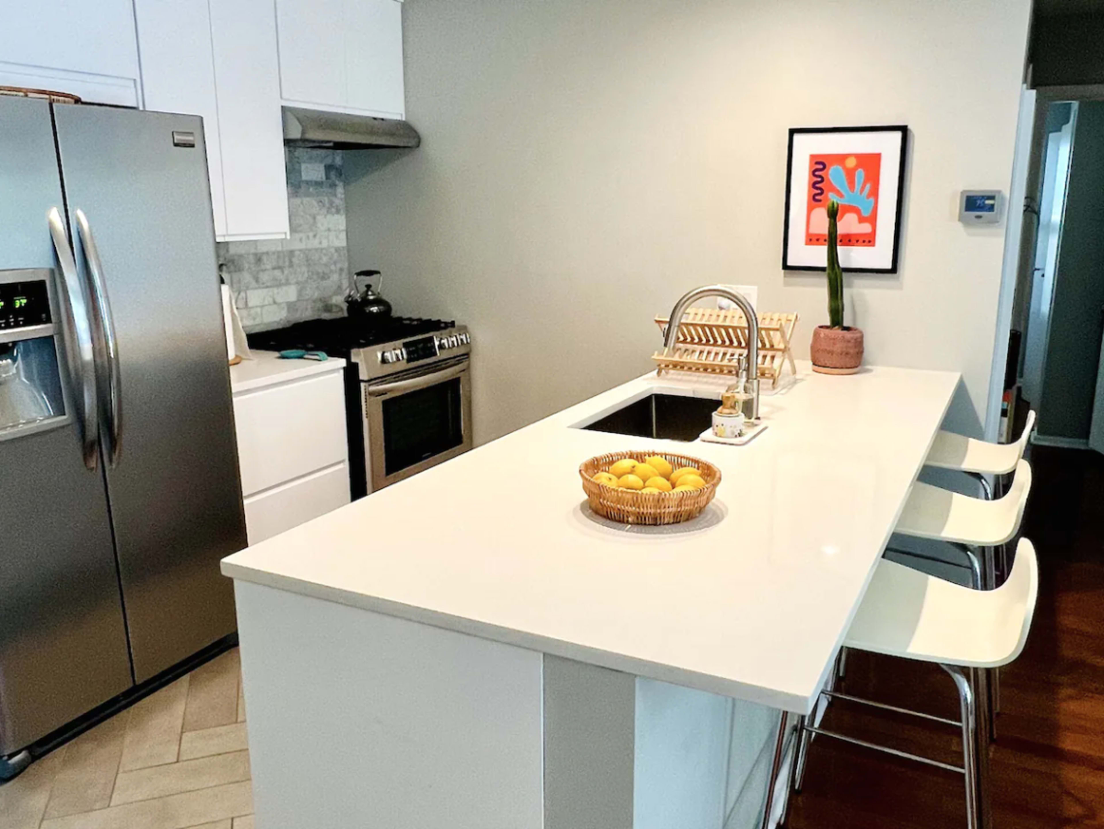 A modern kitchen features a central island with three bar stools, a stainless steel refrigerator, and a bowl of lemons on the countertop.