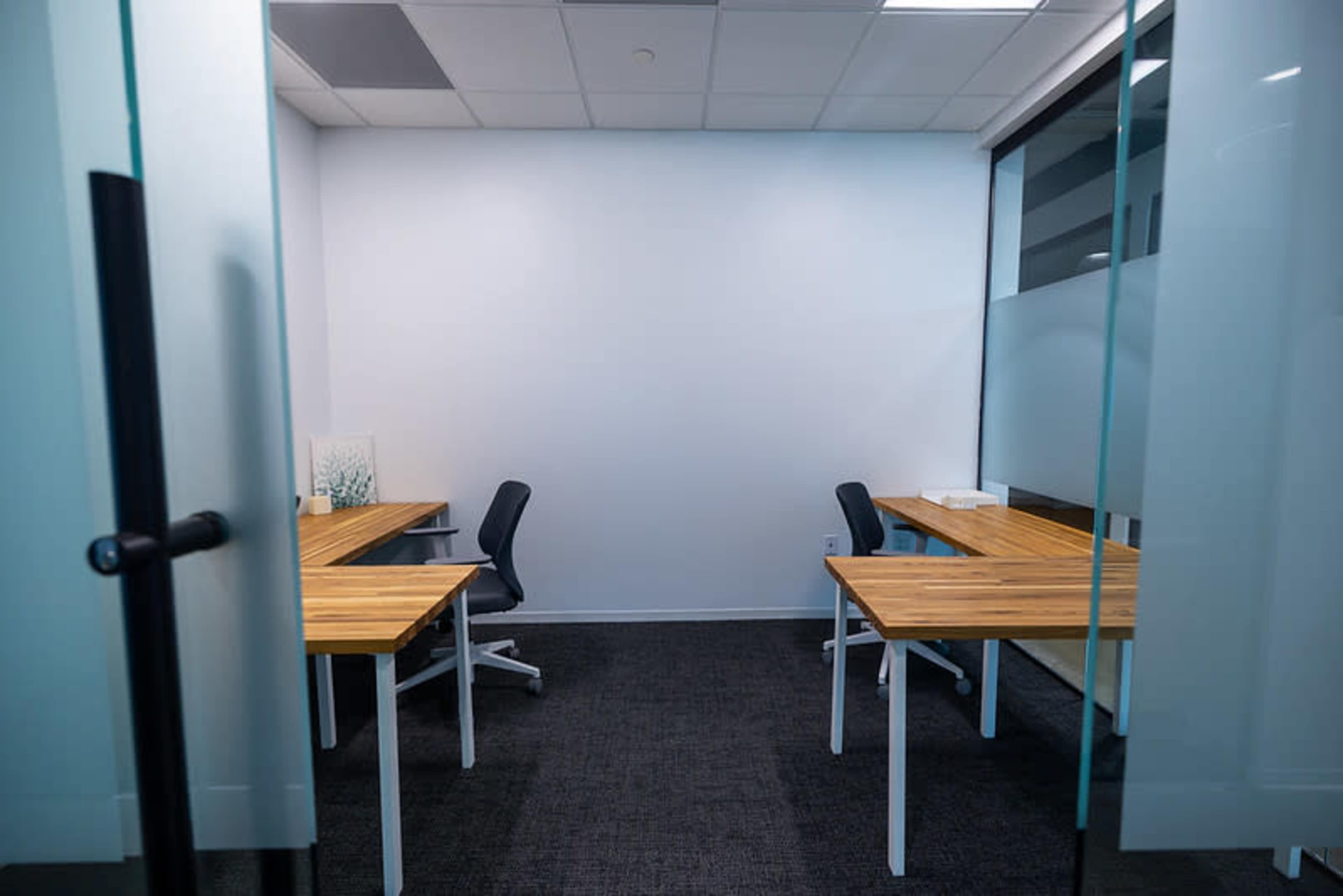 The image shows a small, empty office space with two wooden desks and black office chairs, separated by glass doors.