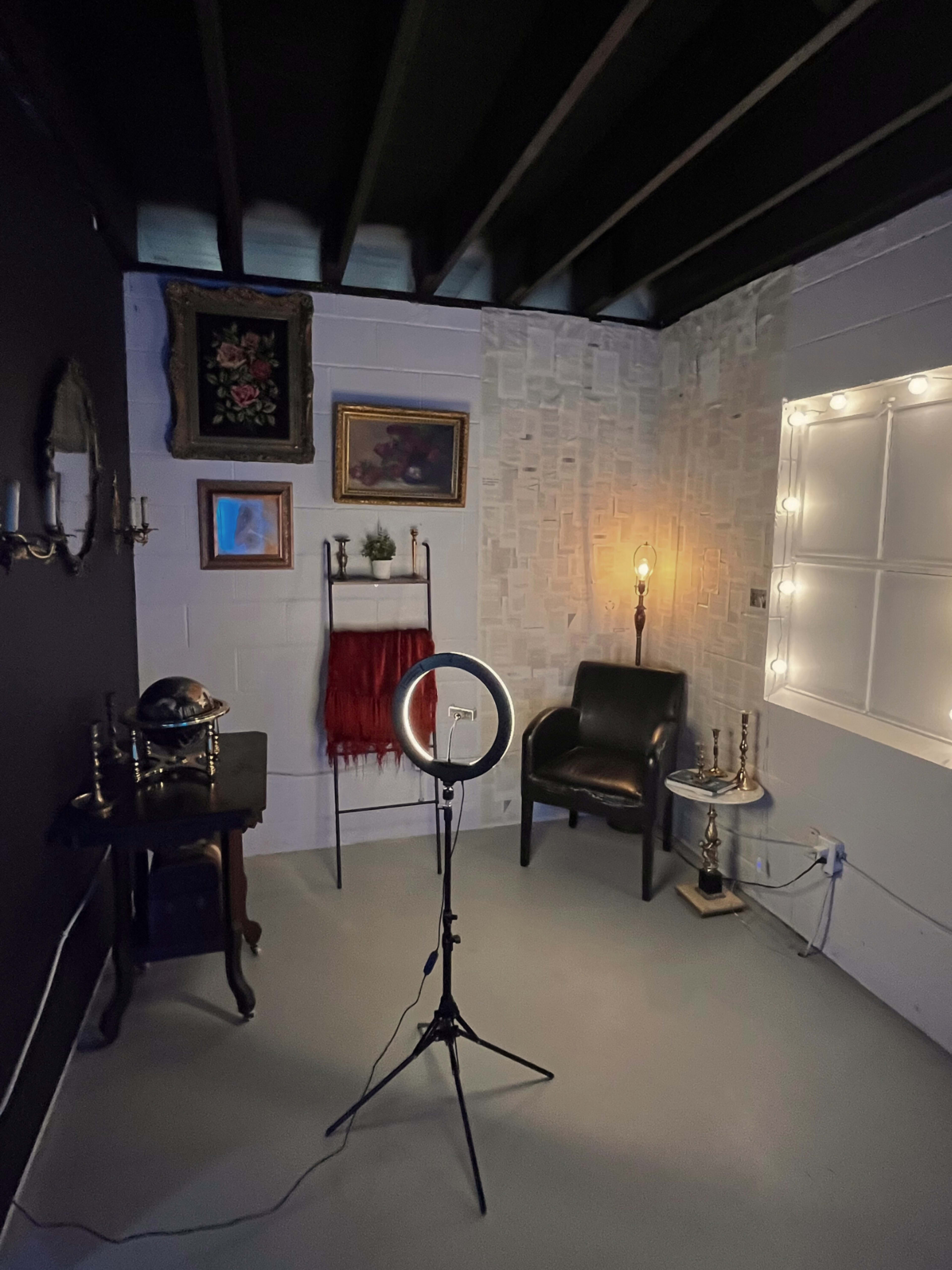 A small, dimly lit room featuring a black wall, assorted artwork, a ring light on a stand, a chair, and decorative items on a shelf.