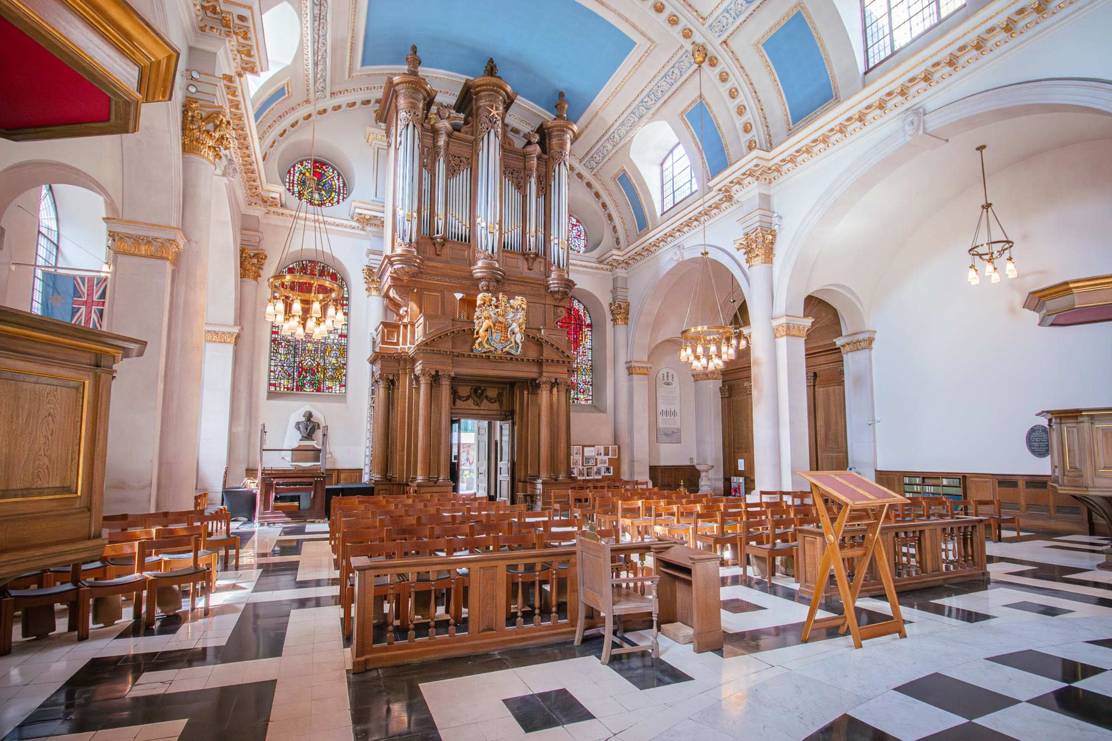 The image shows the interior of a church with a large organ at the front, stained glass windows, and wooden pews arranged on a black and white tiled floor.