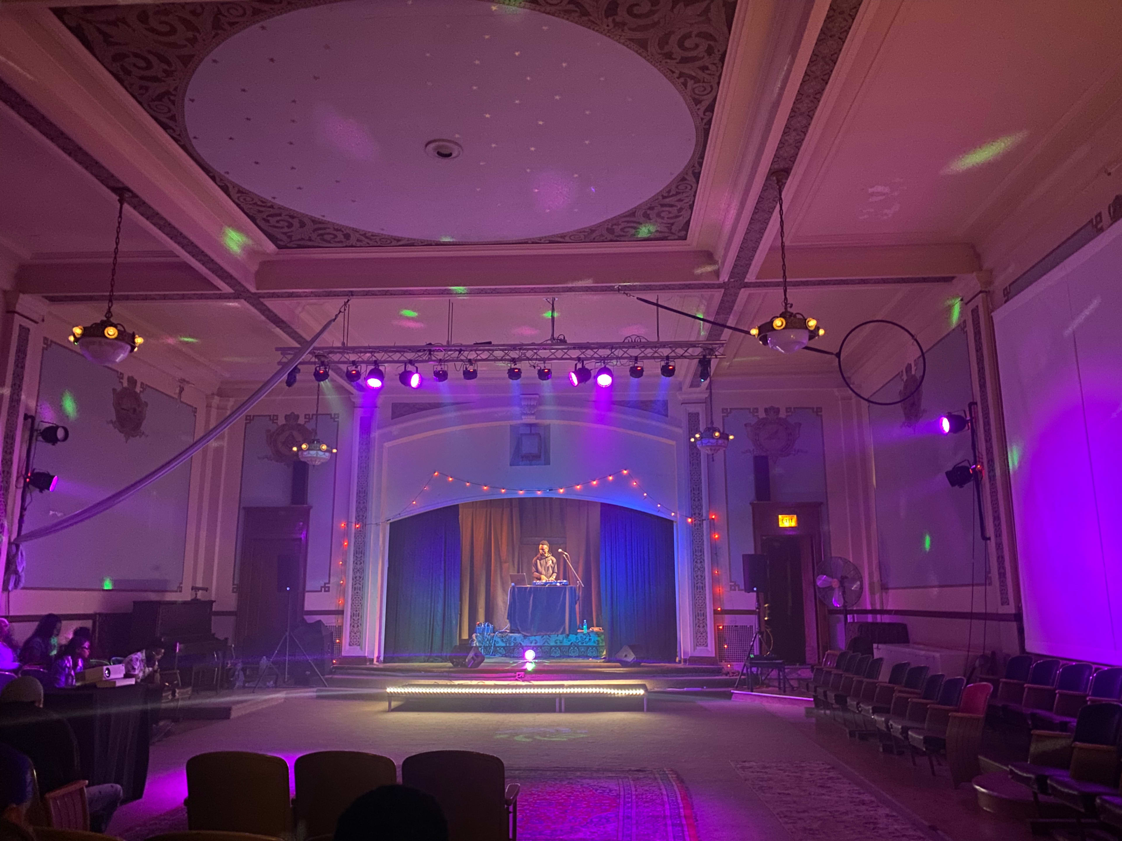 A performer stands on a stage adorned with lights in a decorated hall, surrounded by empty chairs.