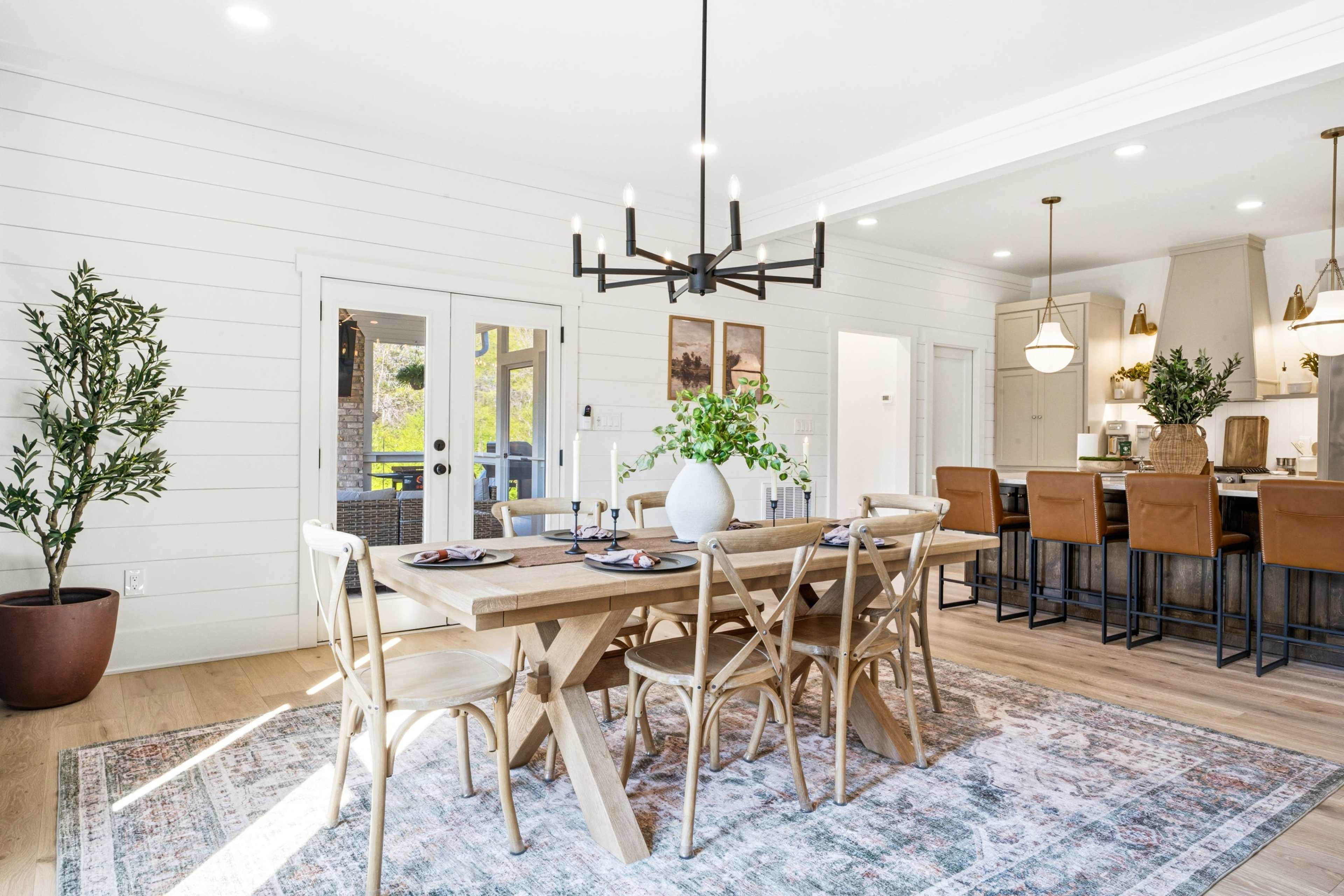 The image shows a modern dining area with a wooden table set for a meal, surrounded by chairs, and an open kitchen area featuring bar stools and decorative elements.