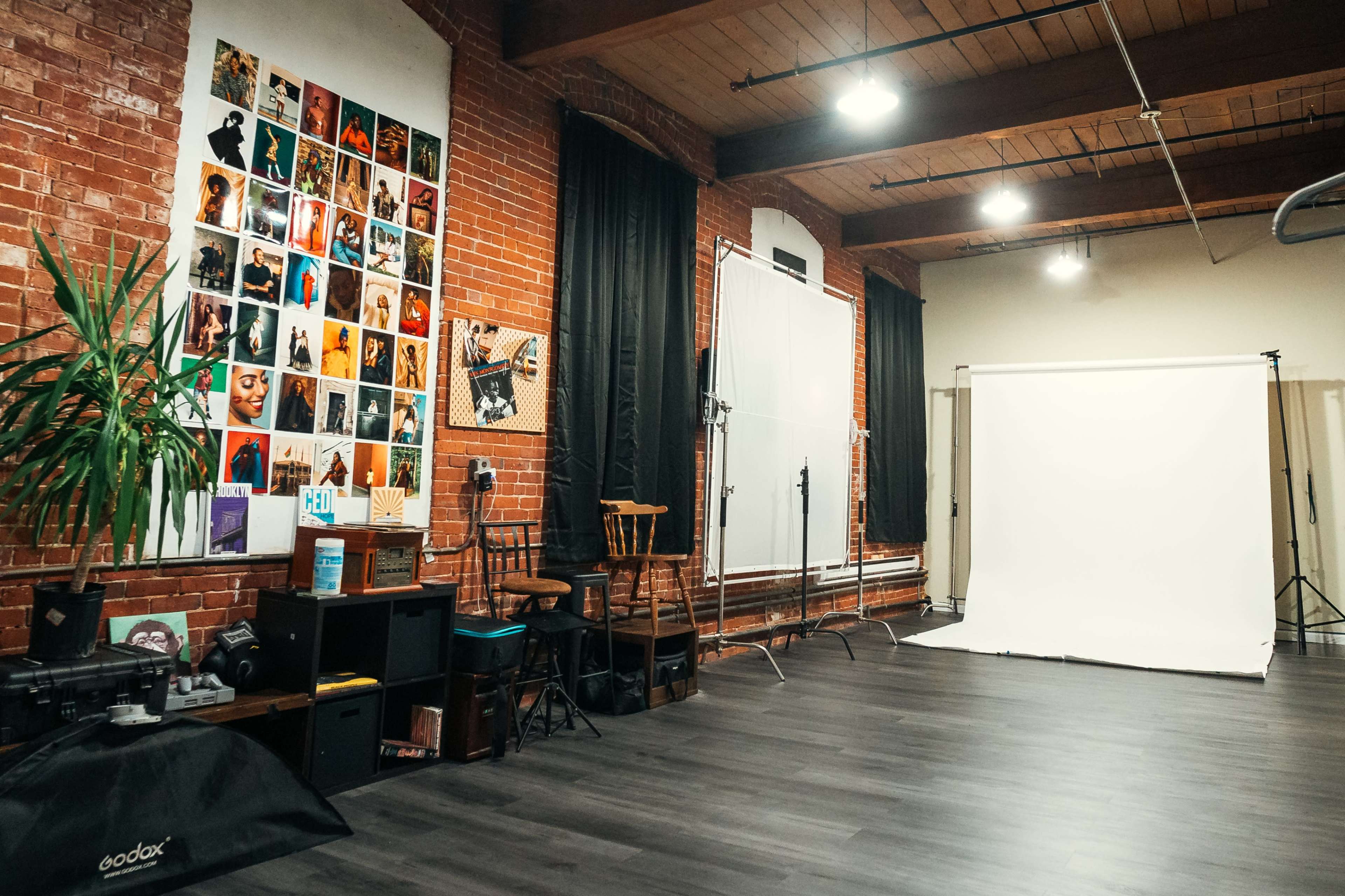 The image shows a photography studio with brick walls, a collection of framed photos, a white backdrop, and various lighting equipment.