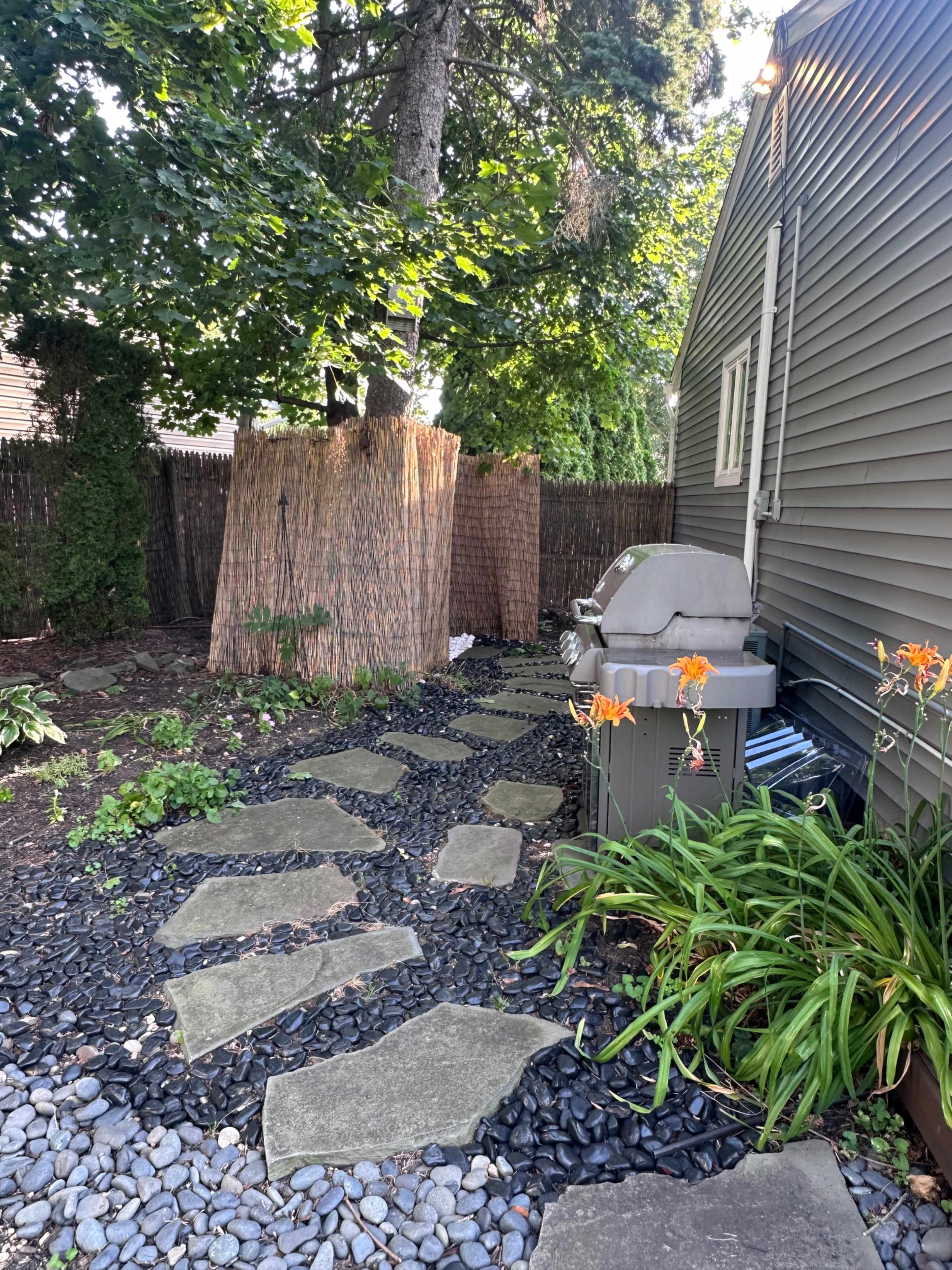 A pathway made of stone slabs leads through a garden area with black pebbles, flanked by tall plants and a grill near a wooden fence.