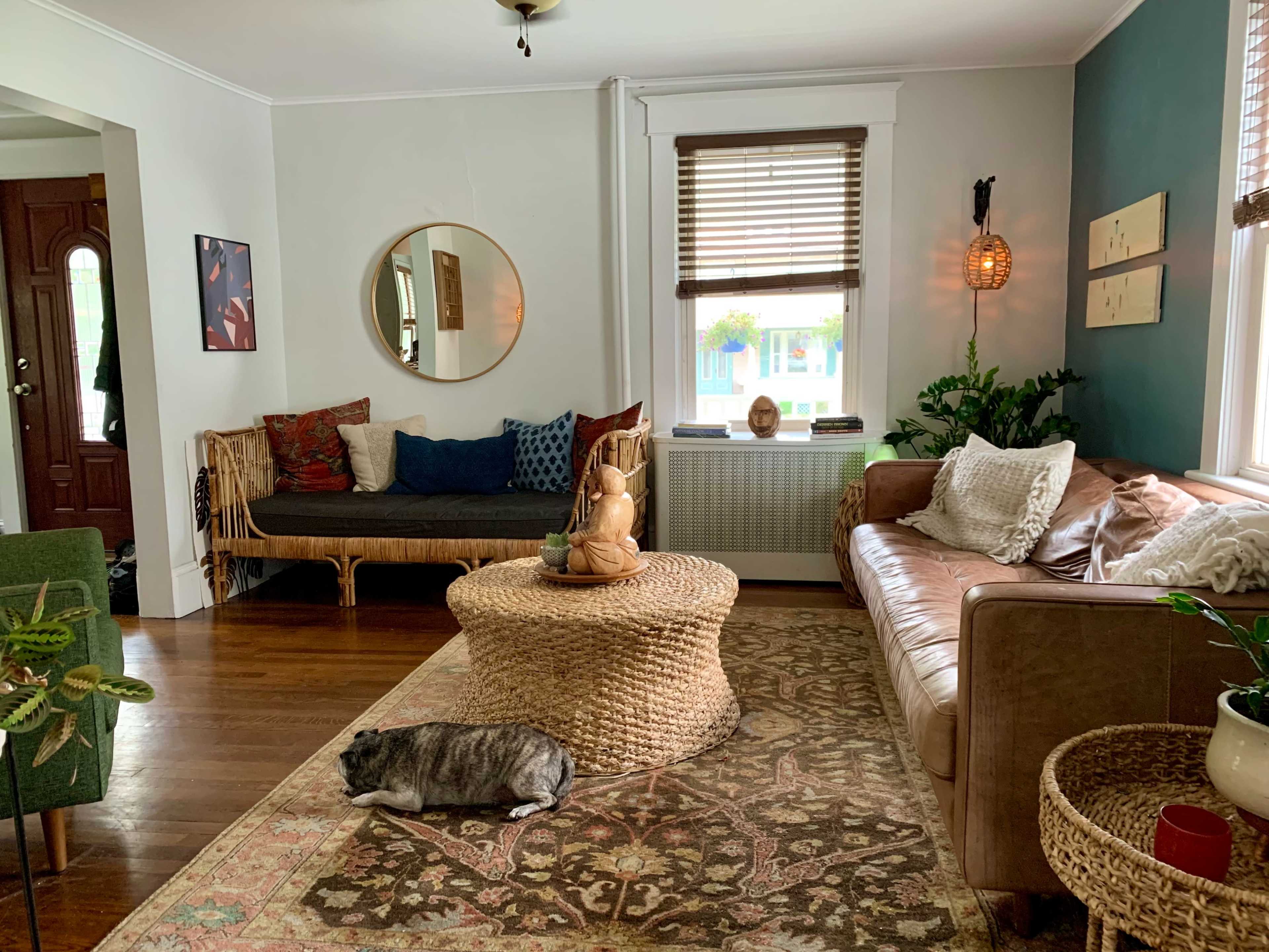 The room features a mix of seating options, including a dark sofa, a leather couch, and a round woven coffee table, with decorative plants and artwork on the walls.