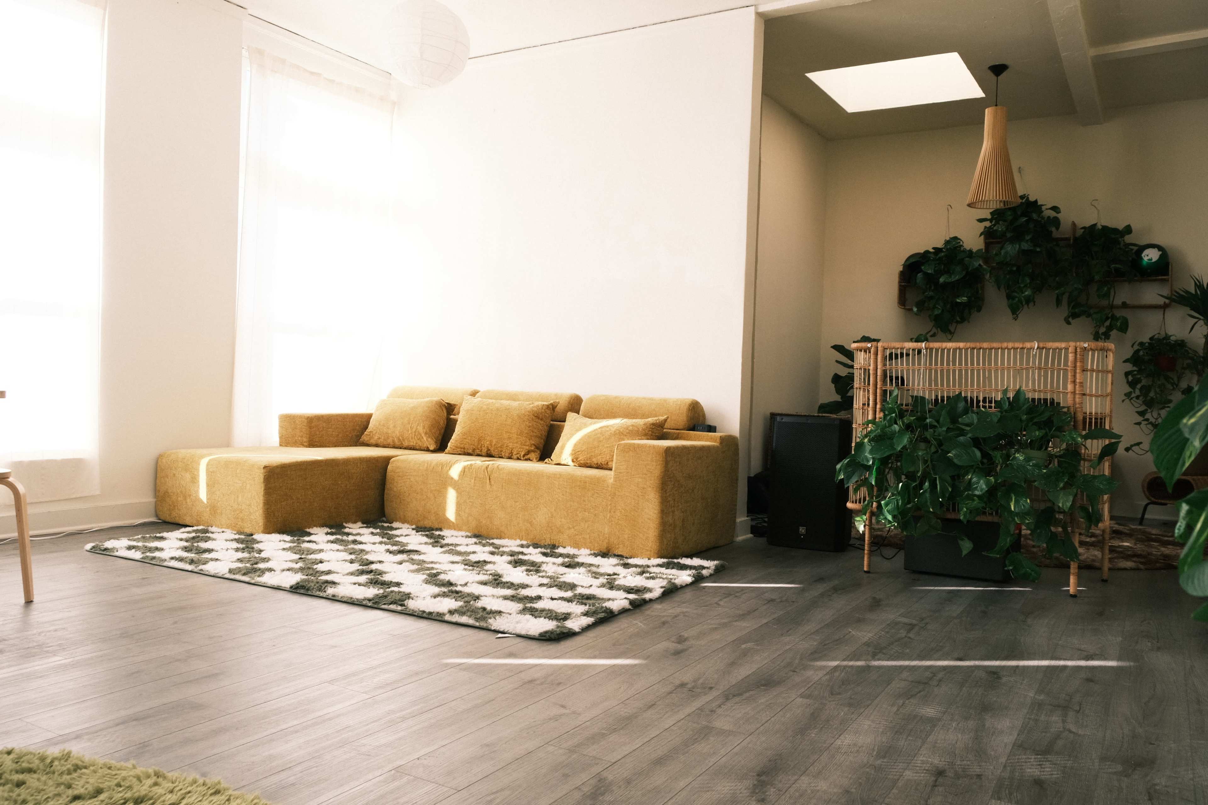 The image shows a bright, minimalist living room featuring a yellow sectional sofa, a patterned rug, and various plants arranged in a well-lit space.