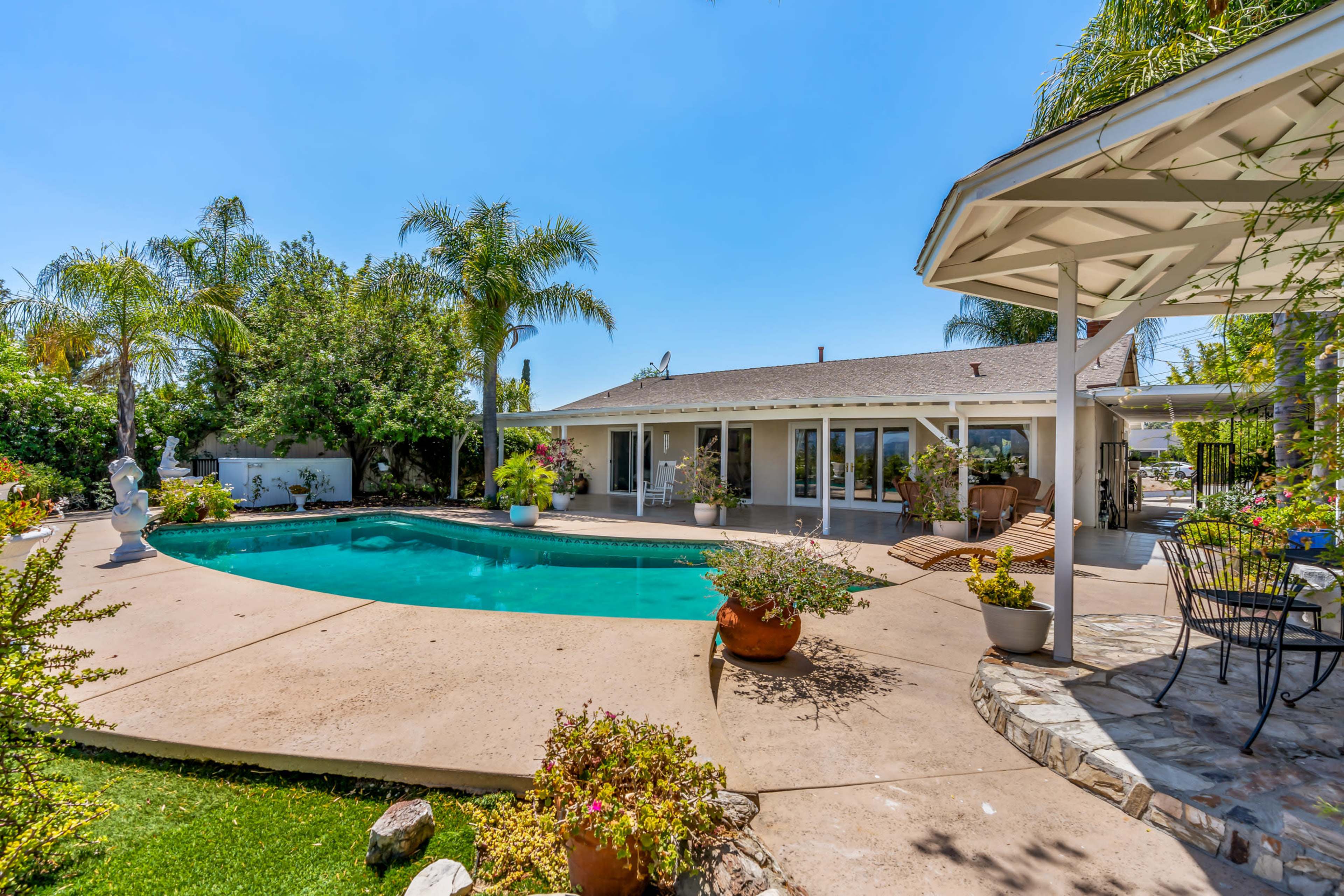 A backyard with a clear swimming pool, surrounded by greenery and a patio with seating areas.