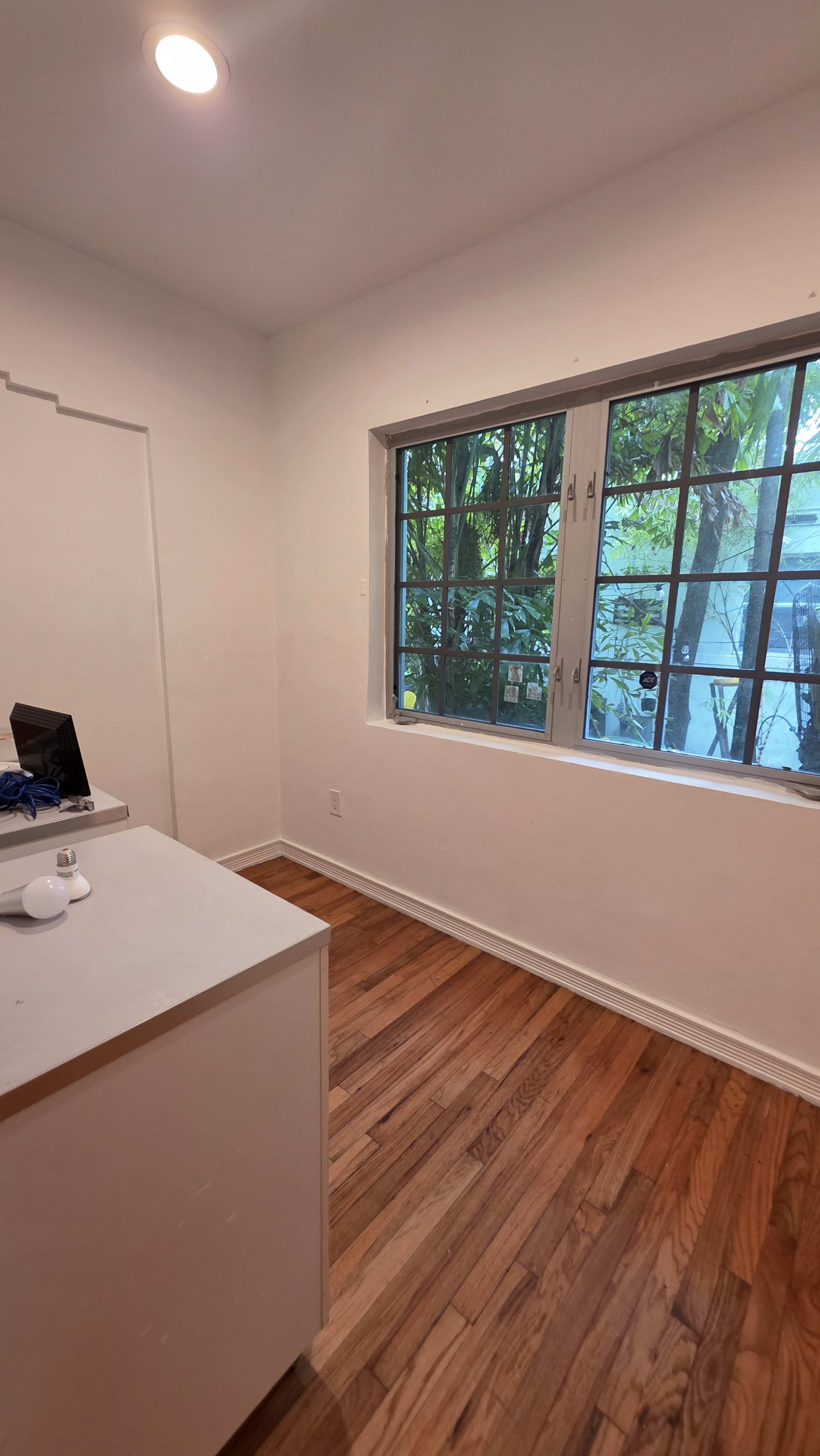 A small room with wooden floors features a window with several panes overlooking greenery and contains a minimal desk setup against one wall.