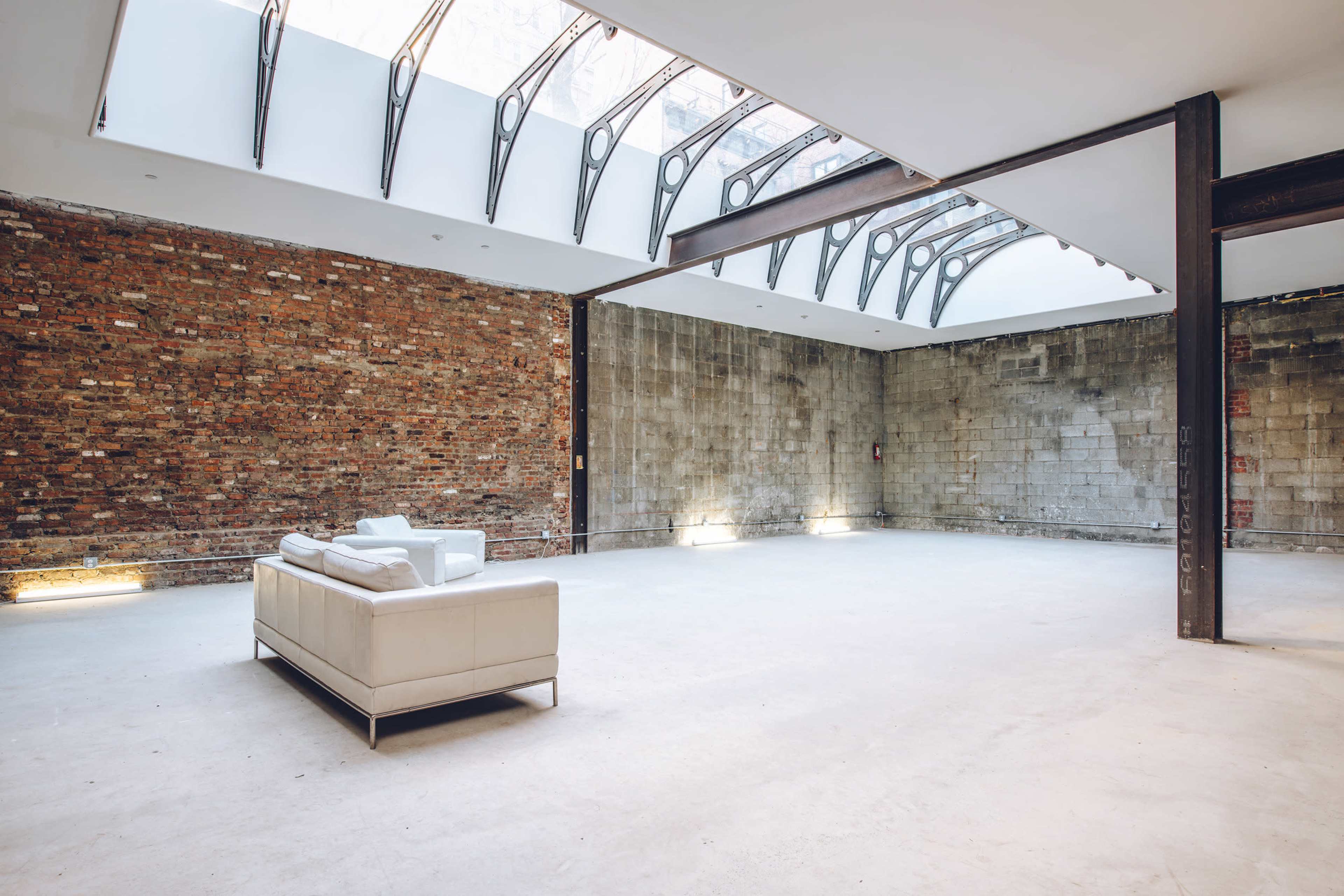 A spacious interior features a white leather couch against a backdrop of exposed brick and concrete walls, illuminated by natural light from a skylight.