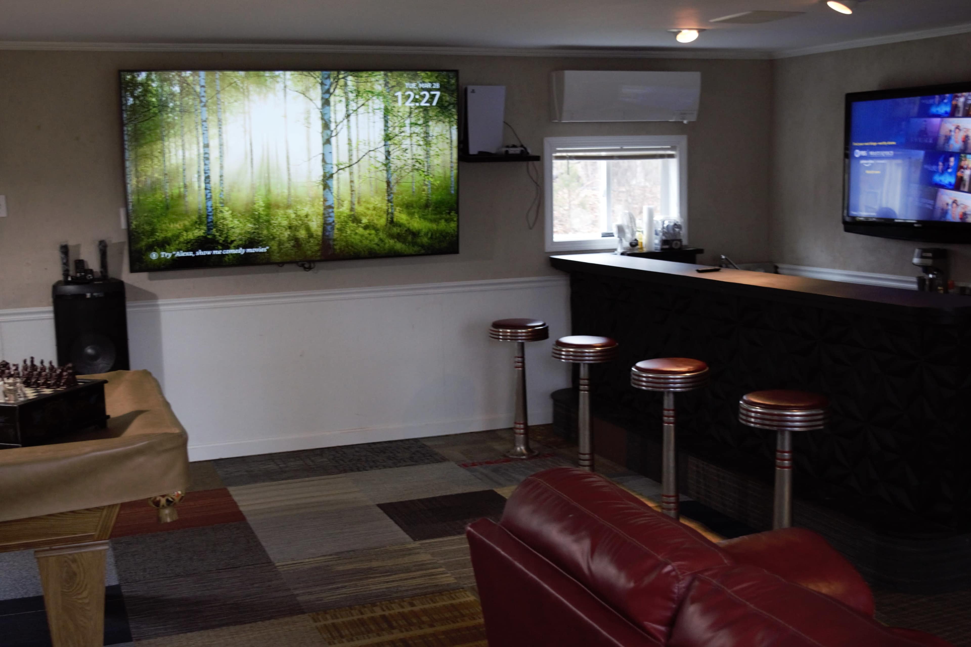 The image shows a room with a large TV displaying a forest scene, a bar area with four stools, and a couch in front of a chess table.