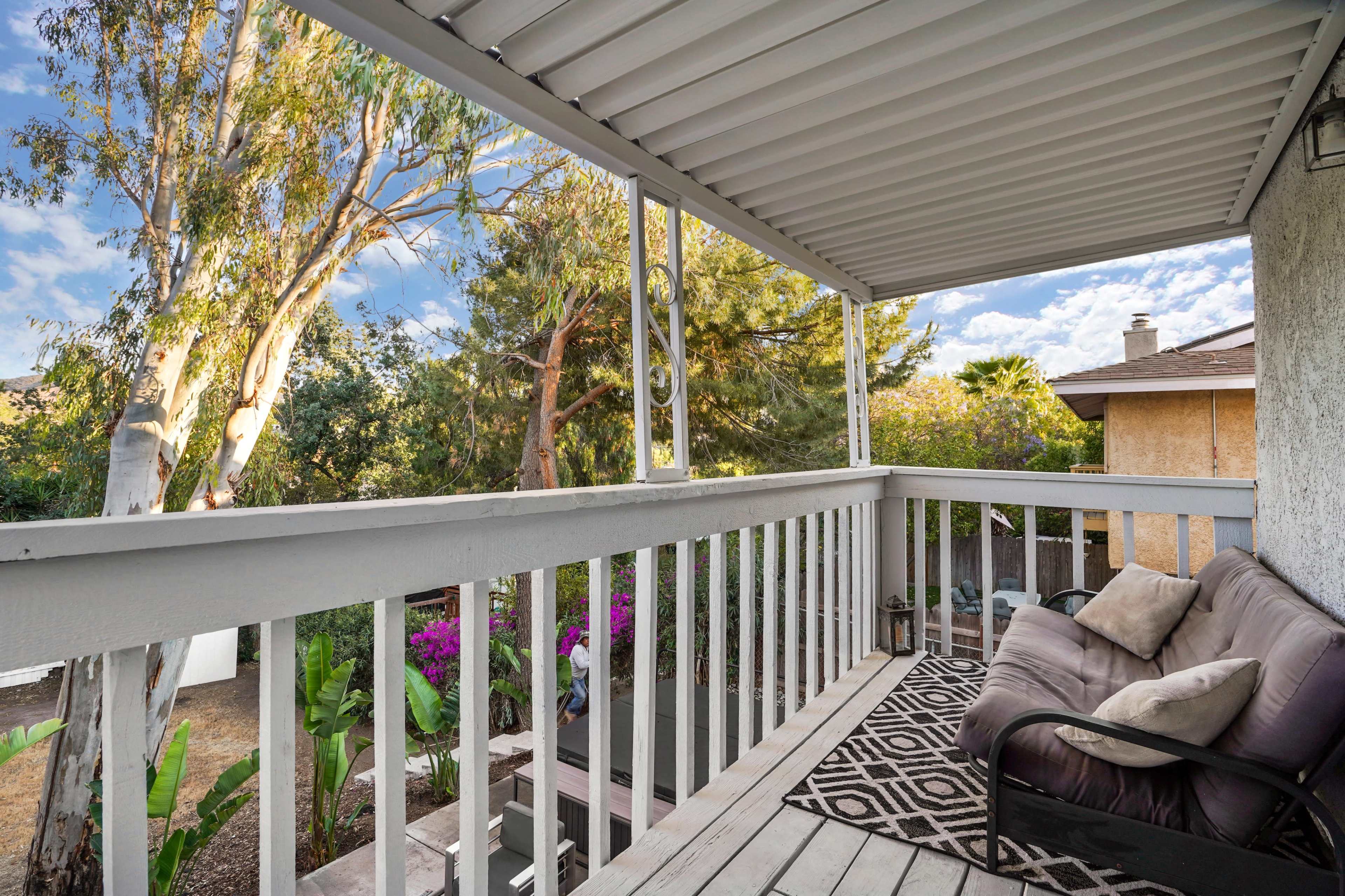The image shows a balcony with a seated area, overlooking a garden with trees and flowering plants.
