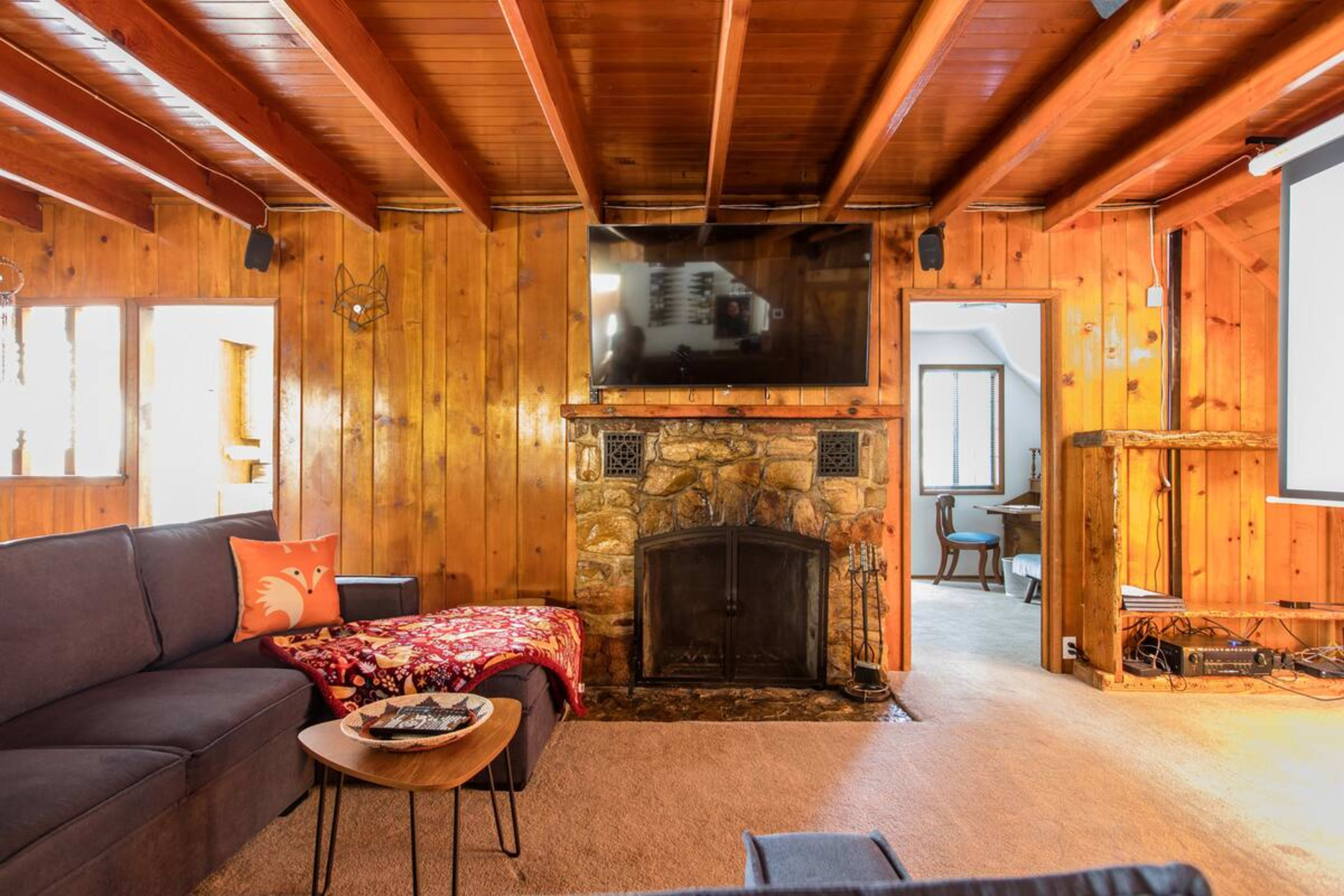 The image shows a cozy living room with wooden walls, a stone fireplace, a large TV mounted above it, and a sectional sofa with a decorative throw and a small coffee table.