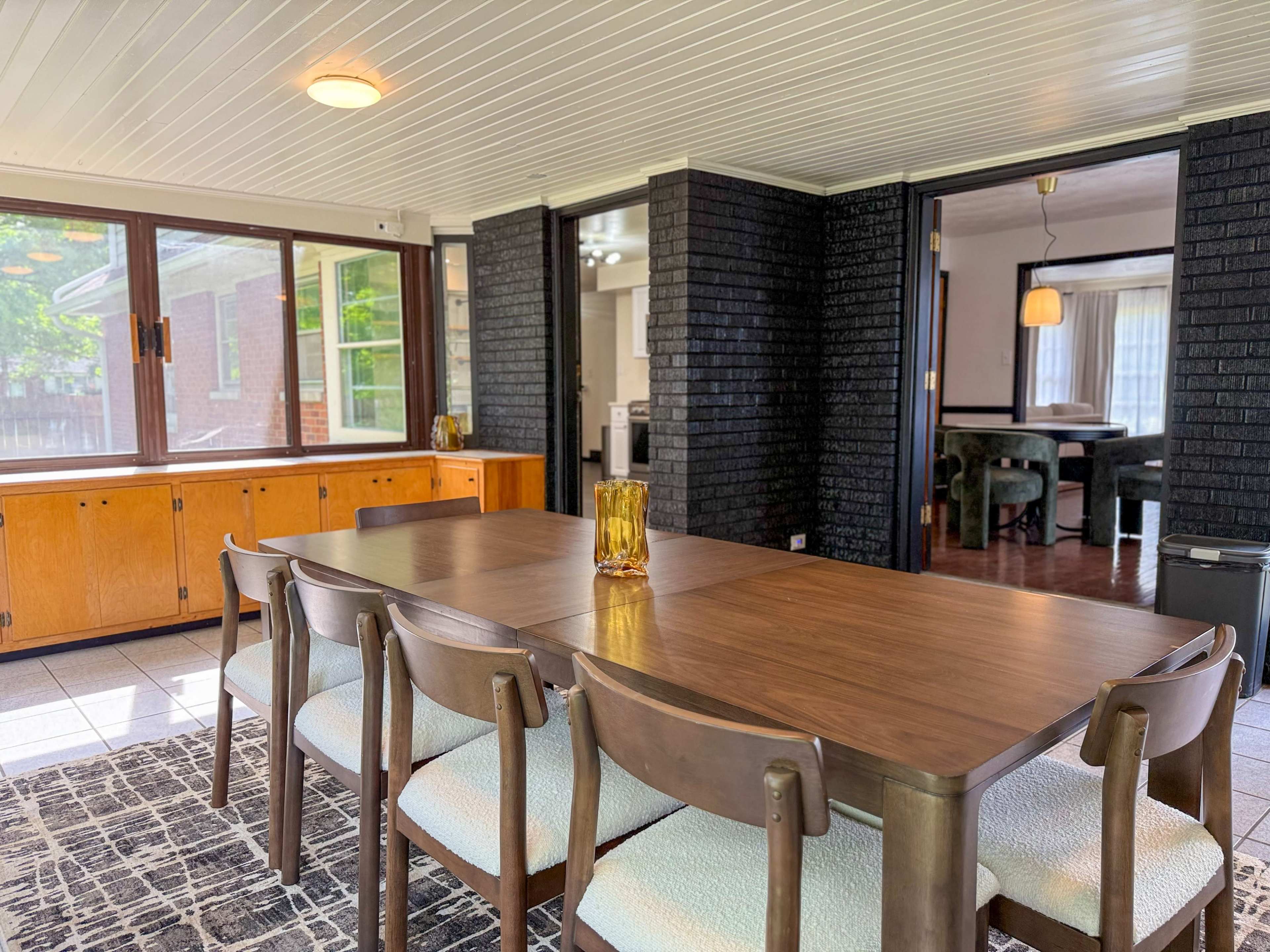 The image shows a dining area with a wooden table surrounded by chairs, adjacent to large windows and an entrance leading to a kitchen space.