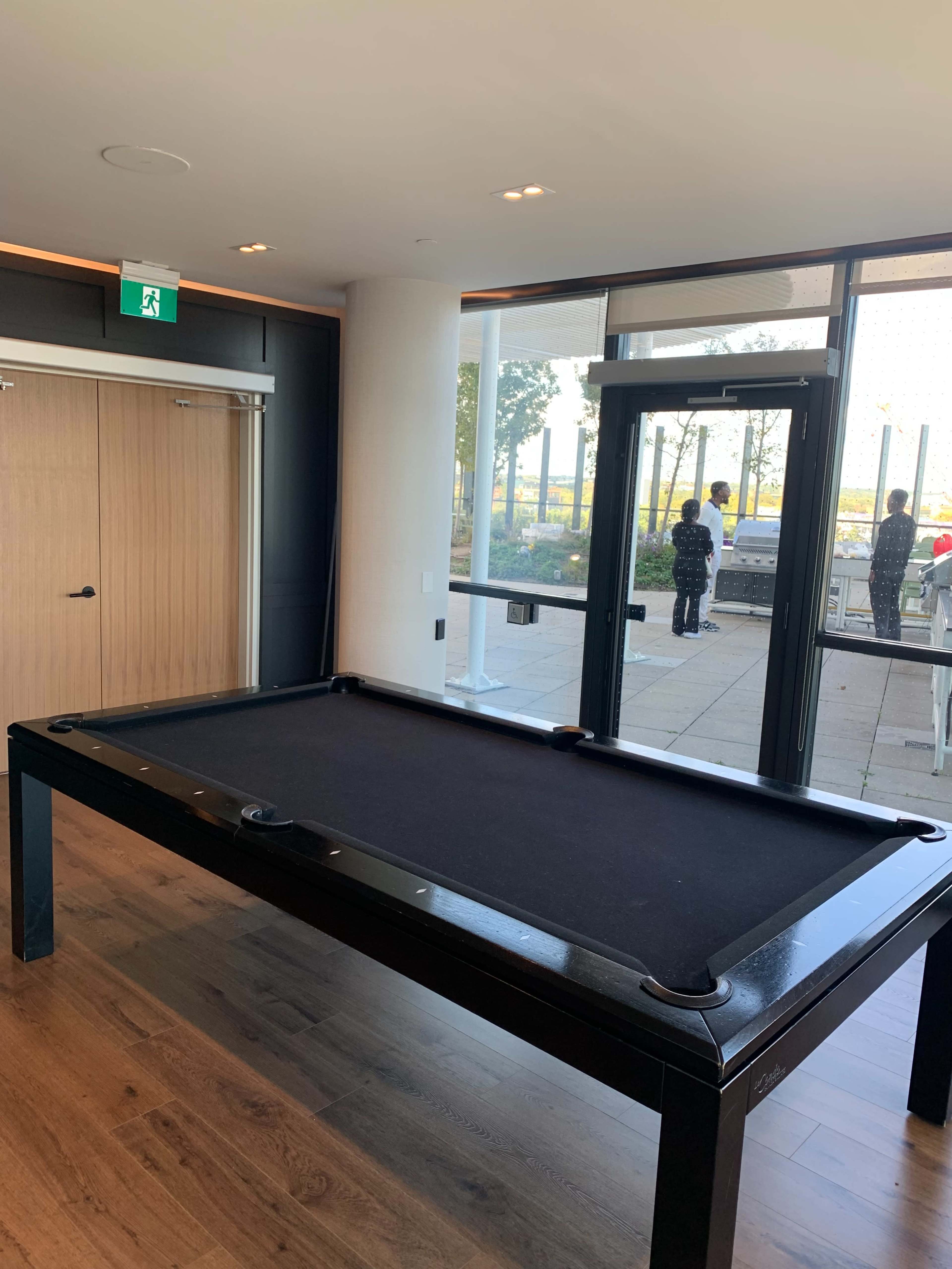 A black pool table is positioned inside a room with large windows and a door leading to an outdoor area.