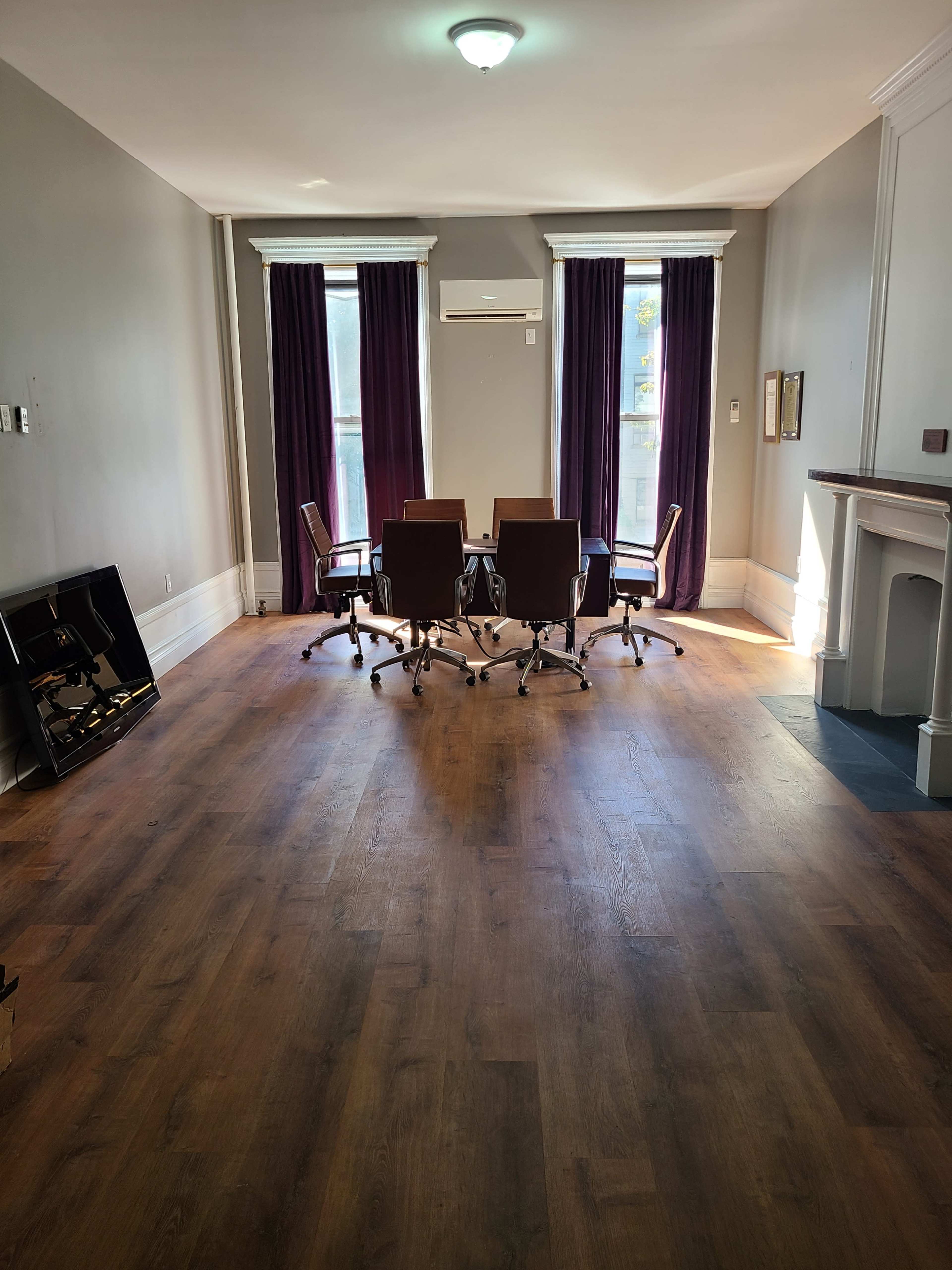 A spacious meeting room features a circular arrangement of five swivel chairs around a central table, with large windows dressed in purple curtains.