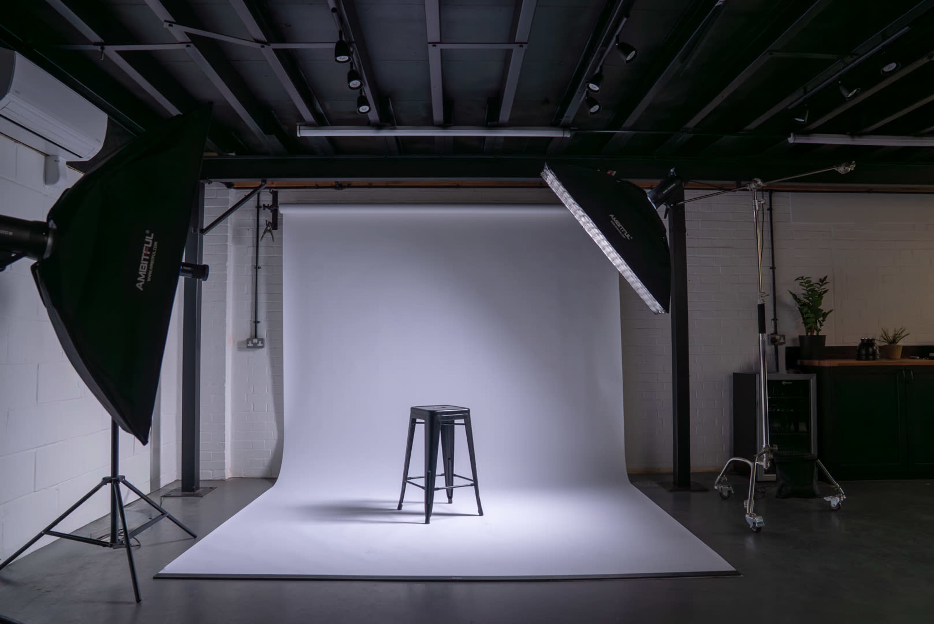 The image shows a photography studio equipped with two softboxes illuminating a black stool on a white backdrop.