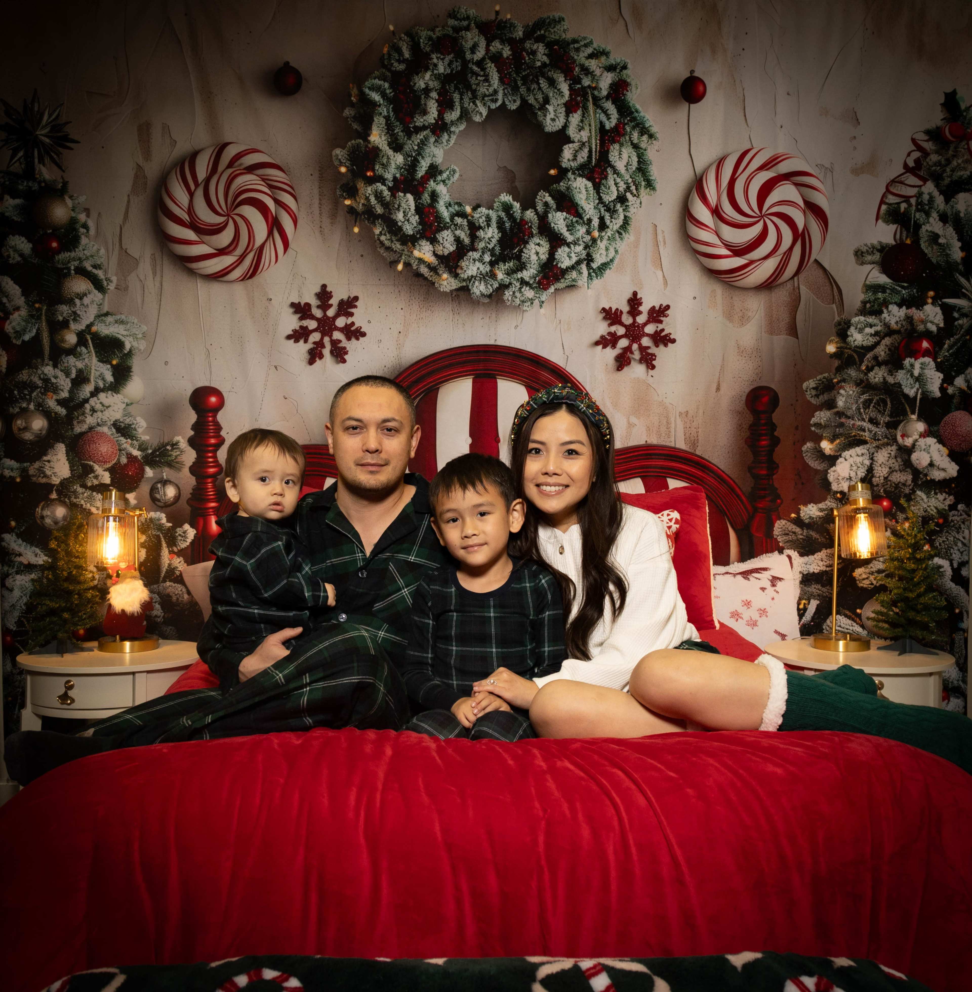 A family of four is seated on a red bed in a festive room decorated with holiday wreaths, candy canes, and Christmas trees.