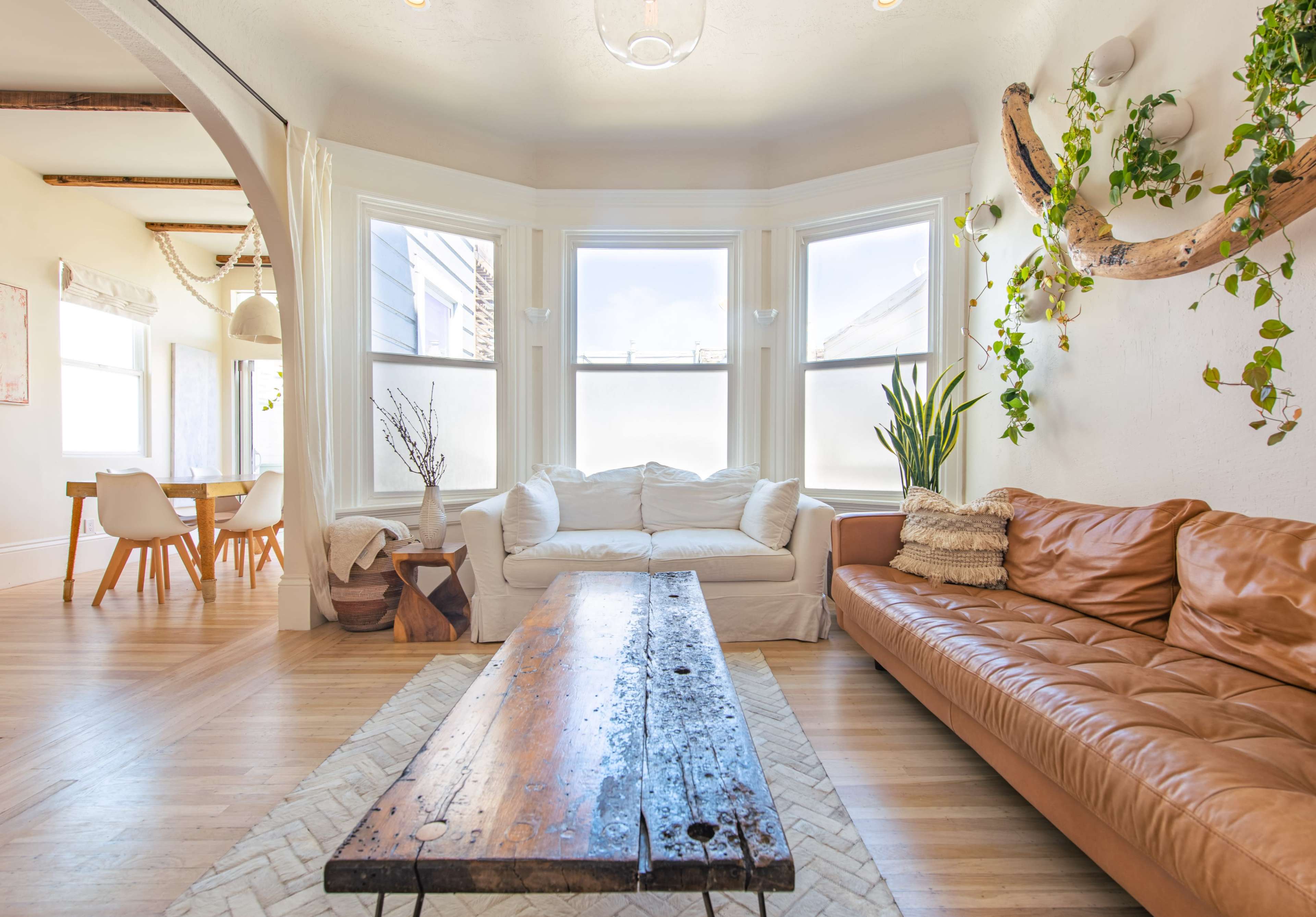 The living room features large windows, a white sectional couch, a brown leather sofa, a wooden coffee table, and potted plants.