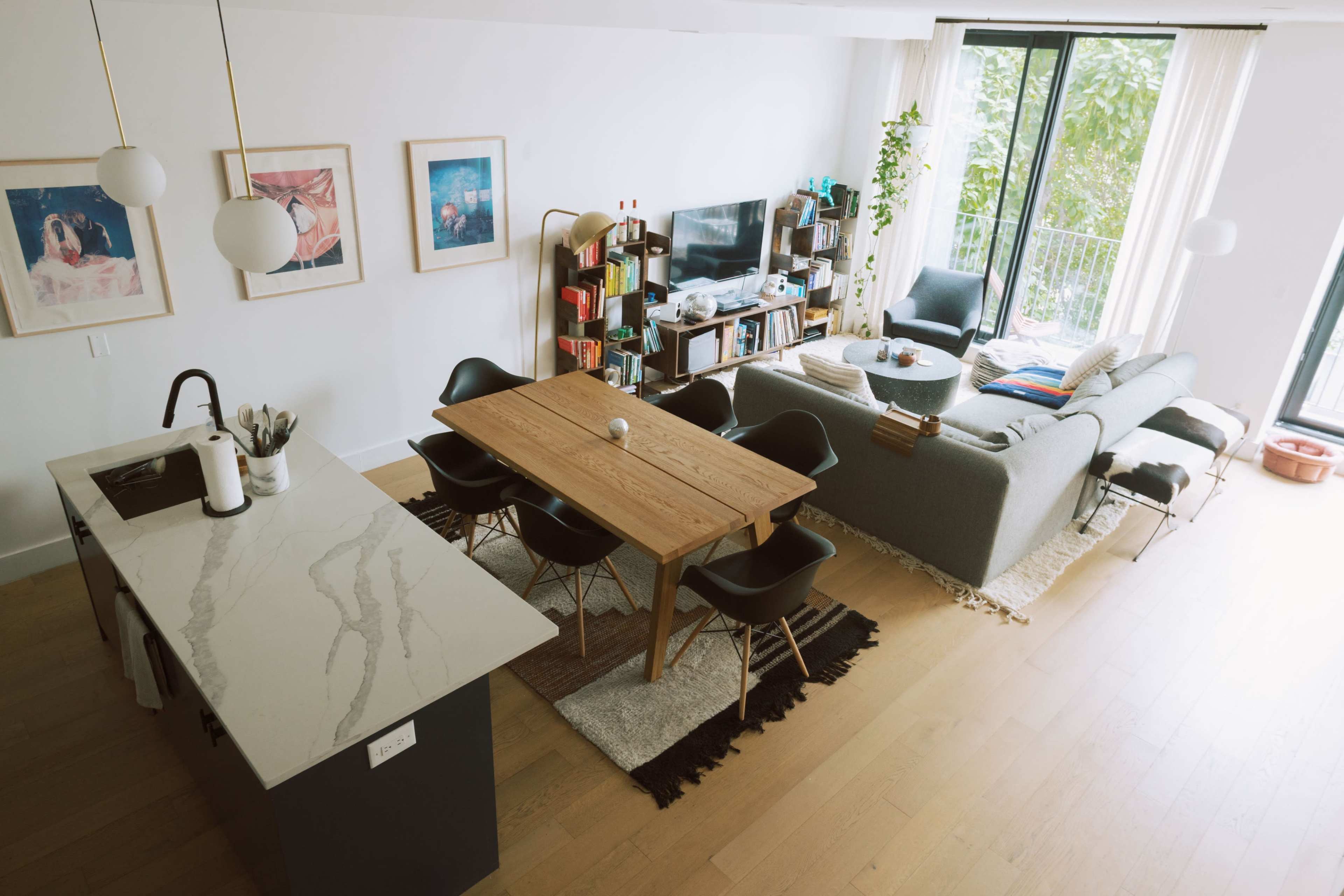 The image shows a modern living room and dining area featuring a wooden dining table, a gray sectional sofa, and a television on a media unit, surrounded by large windows with greenery outside.