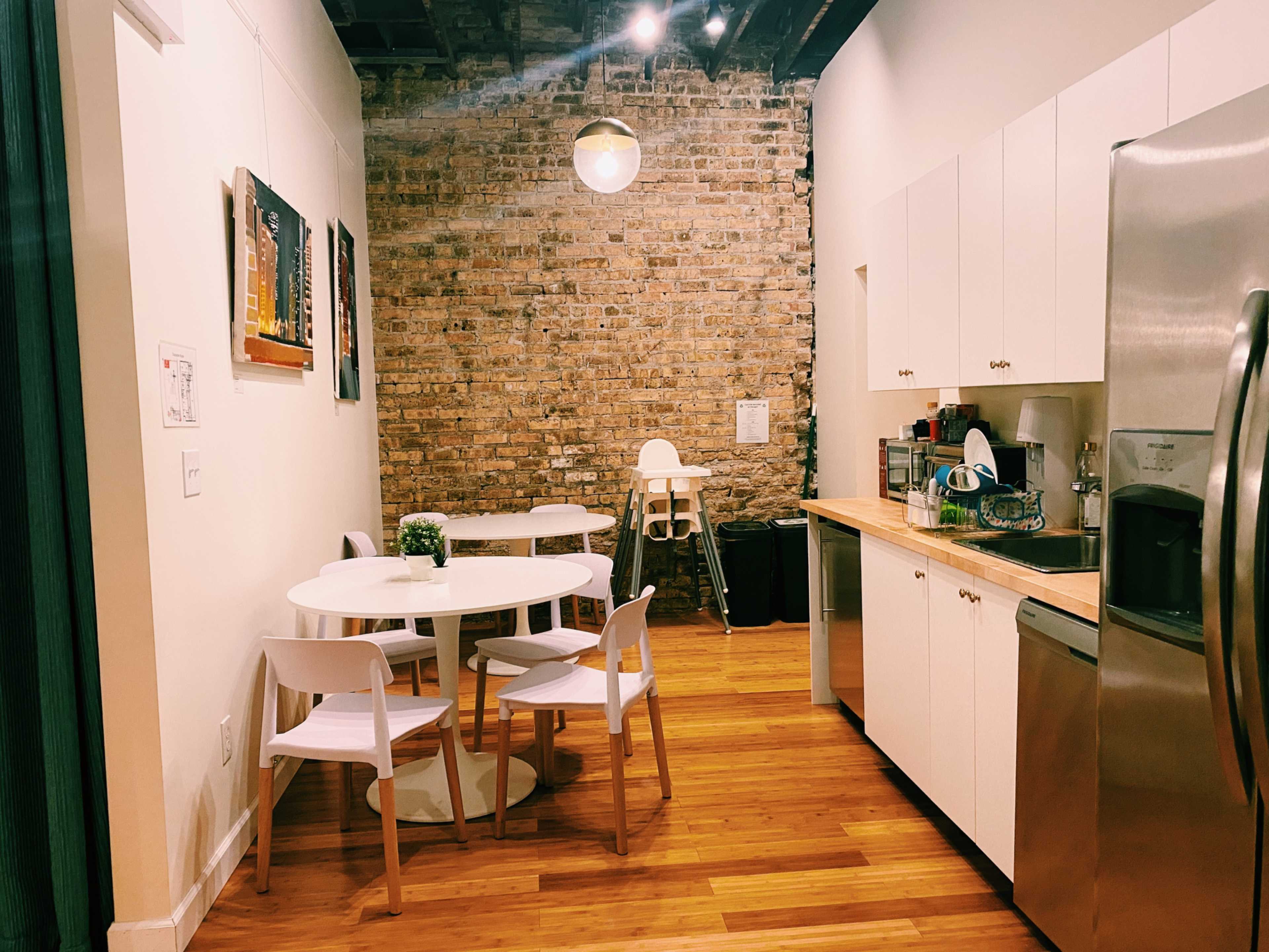 A modern kitchen area with a circular table and white chairs, a wooden countertop, and exposed brick walls.