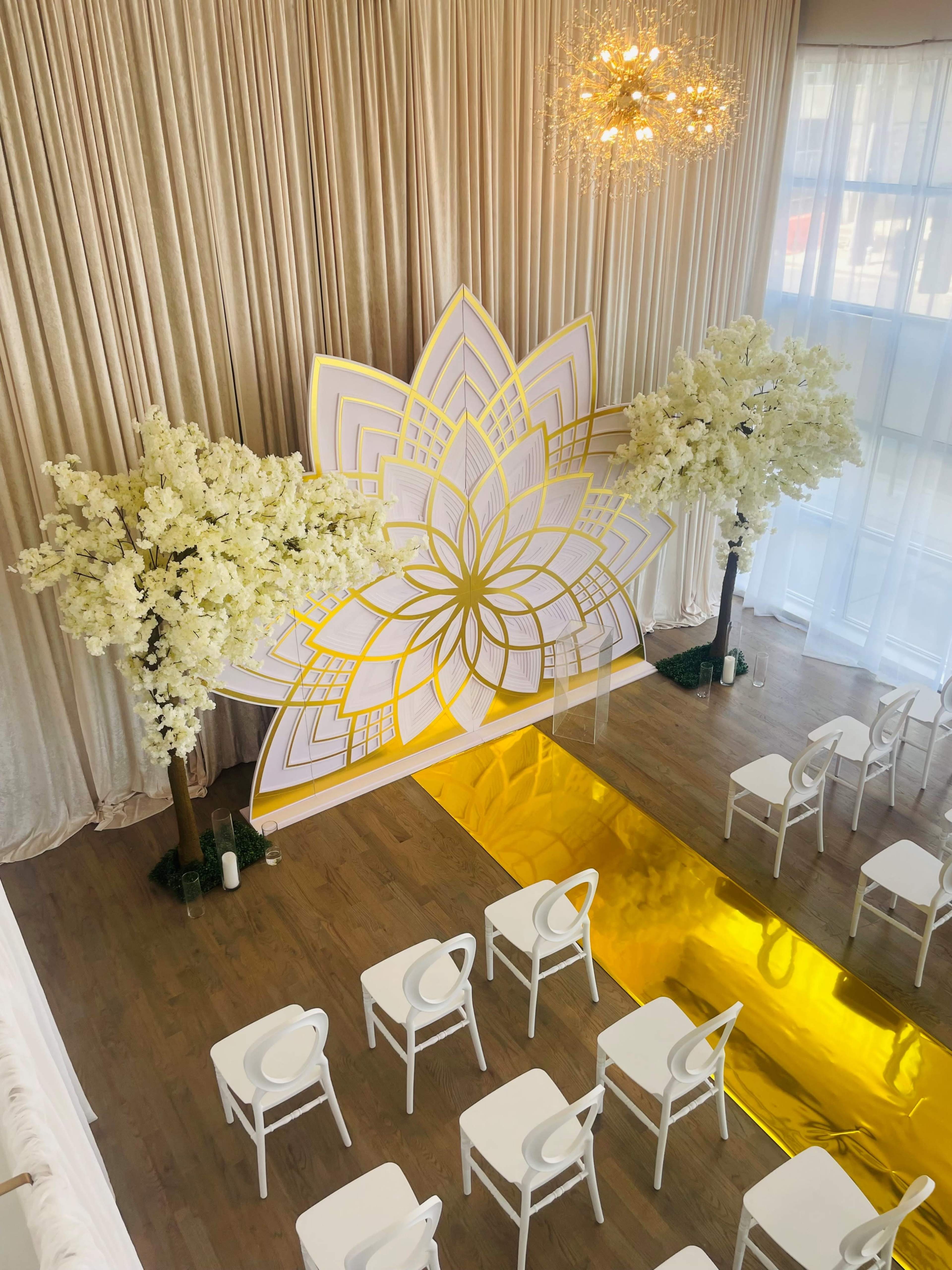 A decorated wedding venue with a large floral backdrop, white chairs arranged in rows, and a golden aisle leading to the stage.