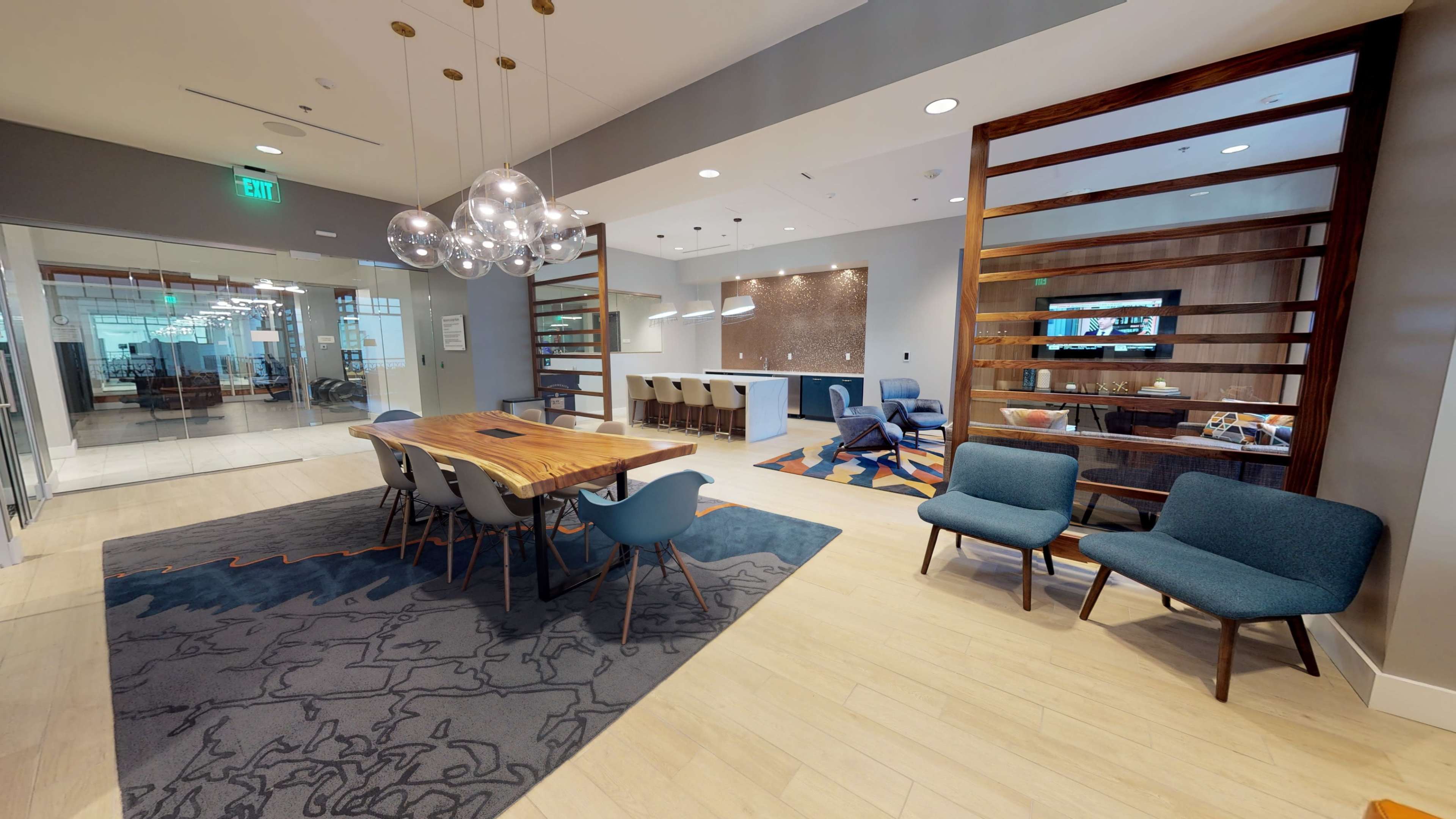 The image shows a modern office space with a dining table surrounded by chairs, a lounge area with blue armchairs, and a well-lit kitchen area in the background.