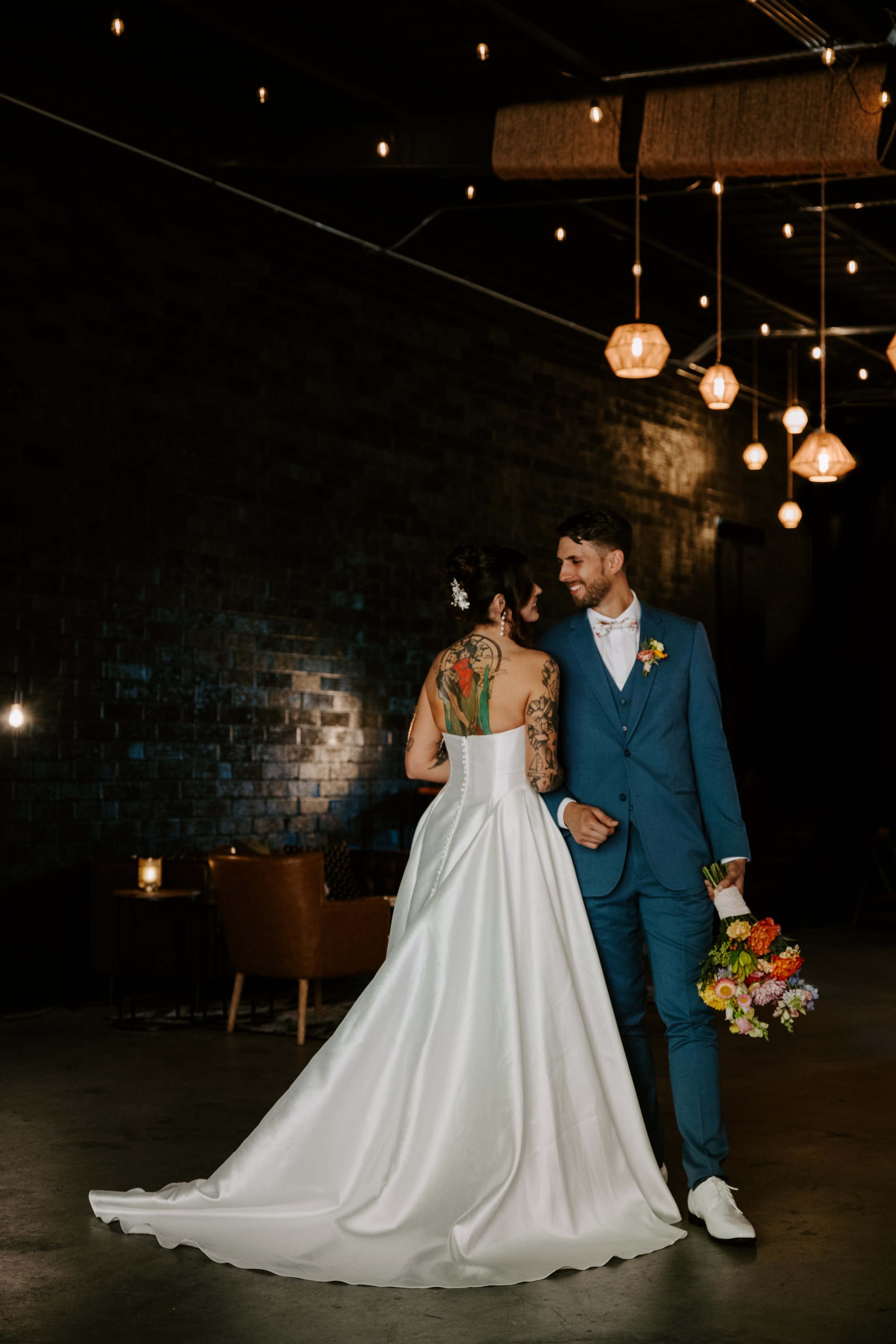 A bride in a white gown with a tattoo on her back stands beside a groom in a blue suit, both facing each other in a dimly lit venue with hanging lights.