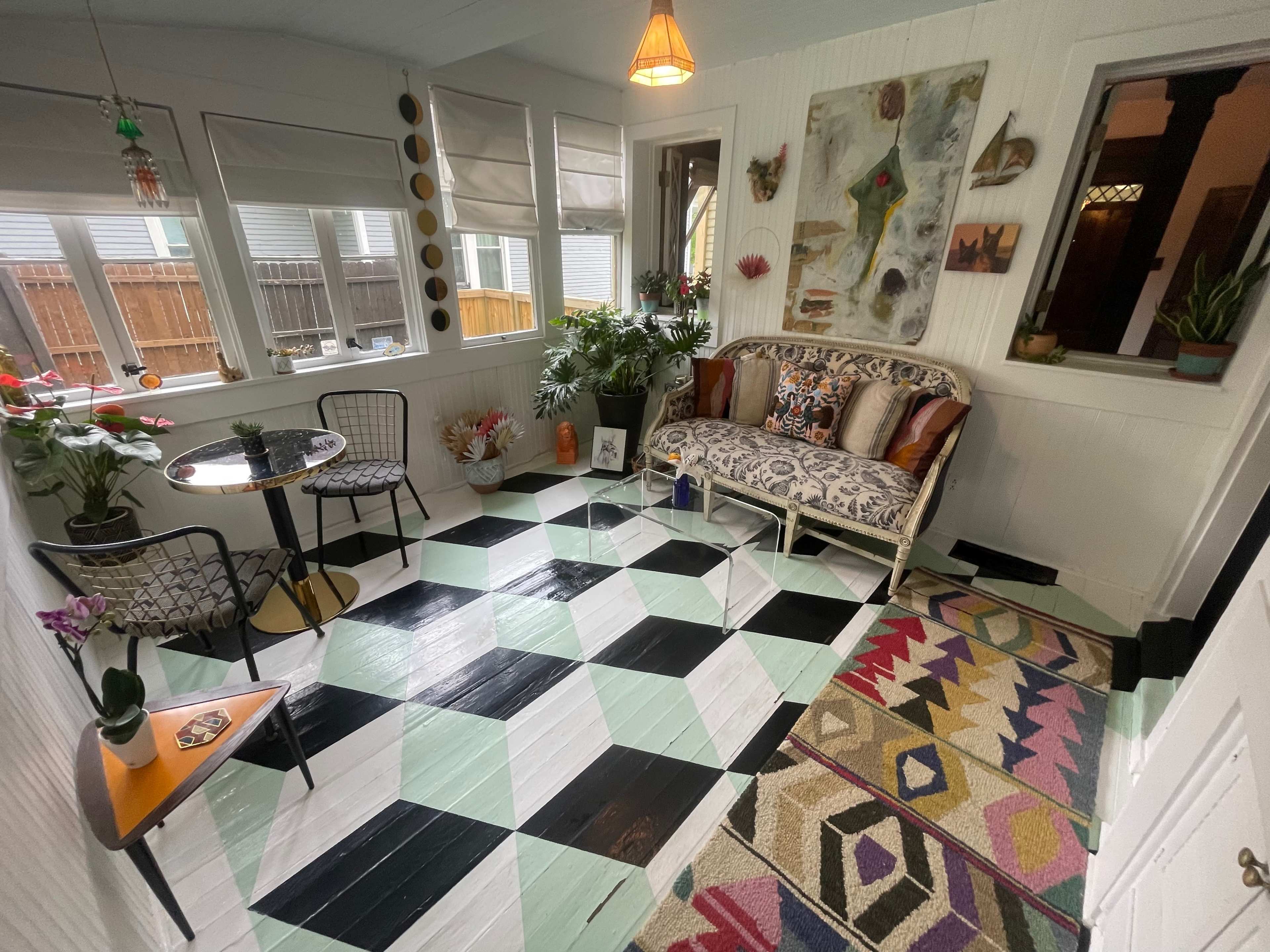 A sunroom with a patterned floor, a patterned sofa, a small table with chairs, and various plants and artwork on the walls.