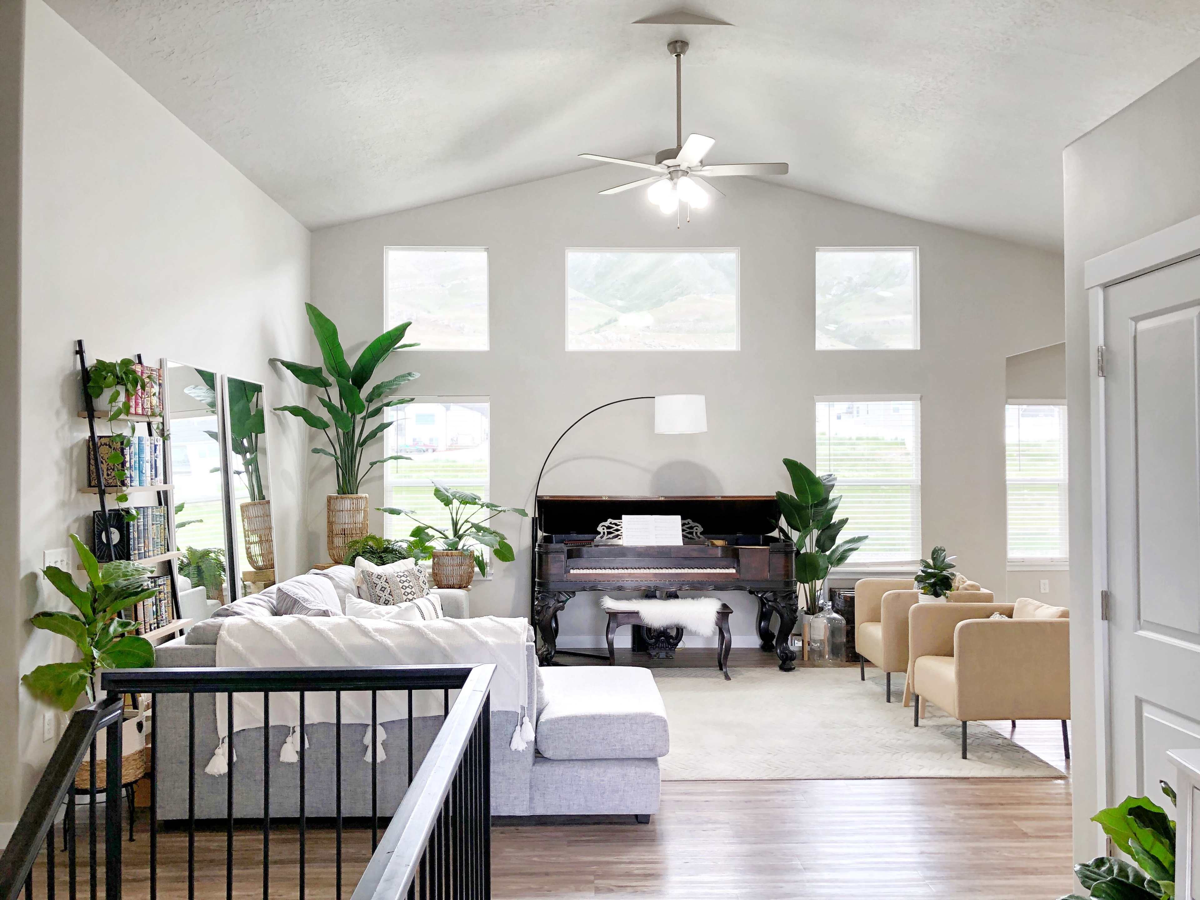 A spacious living room featuring a plush sectional sofa, a piano, two armchairs, and large windows that let in natural light, all adorned with various indoor plants.