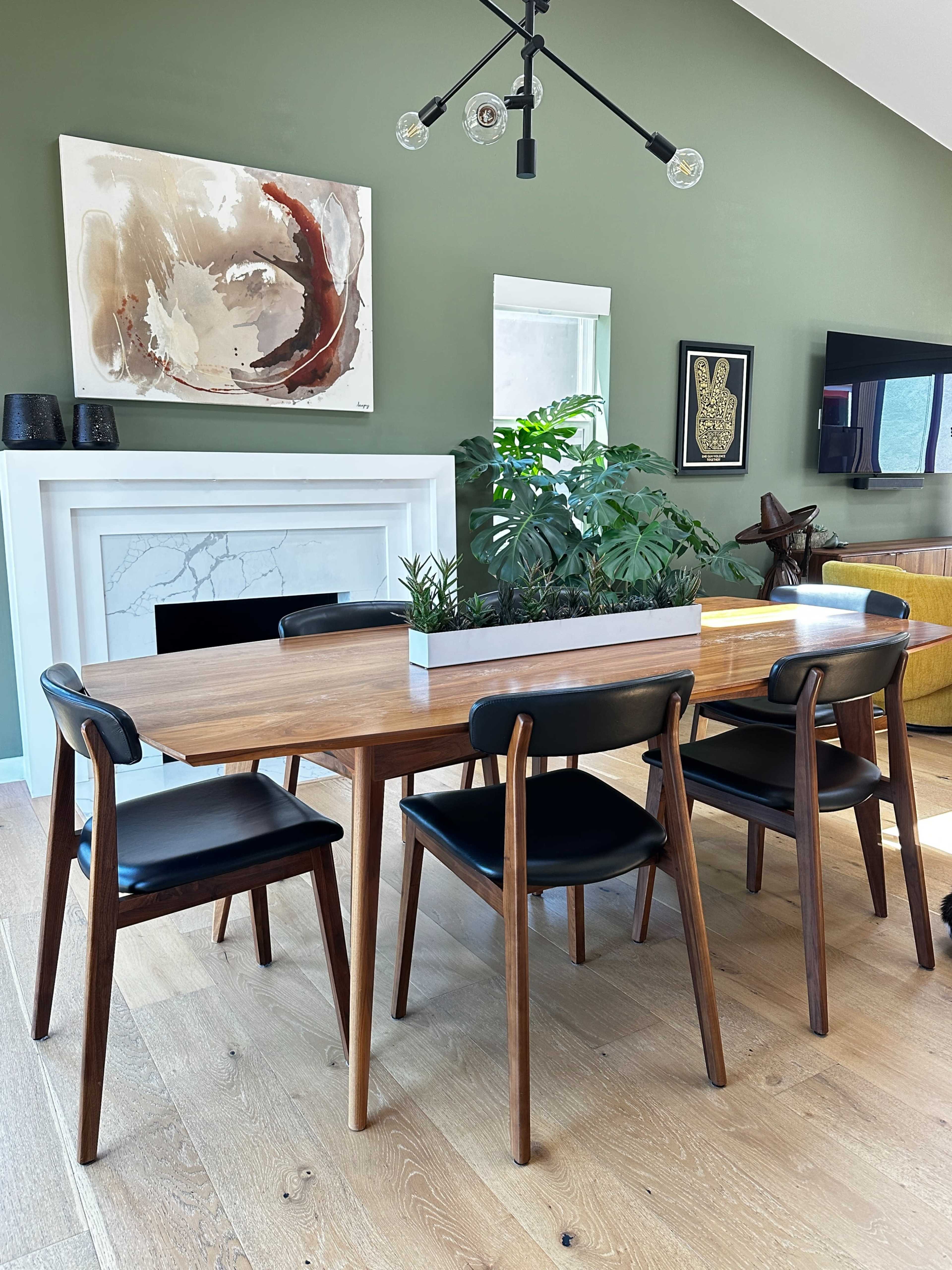 A wooden dining table with black chairs is arranged in a room featuring a fireplace and green walls adorned with abstract art and decorative elements.