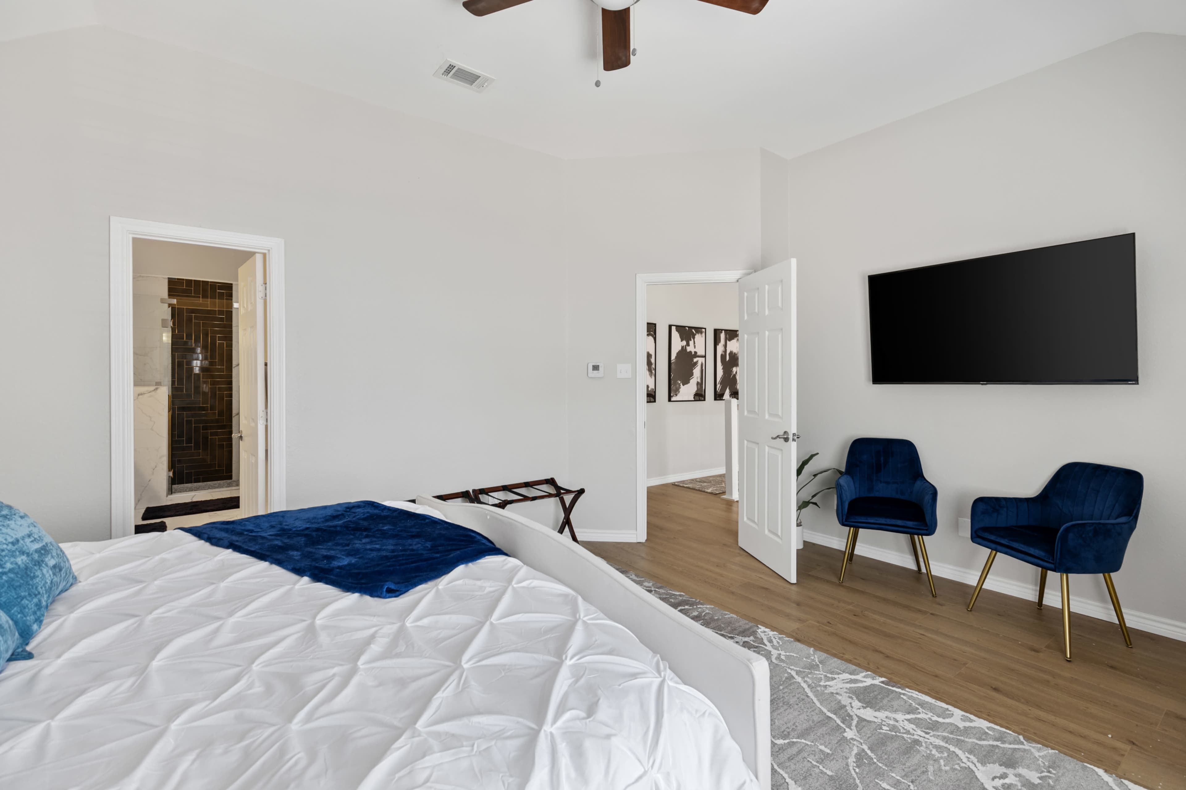 A modern bedroom features a large bed with a blue throw, a wall-mounted television, and two chairs in a neutral-toned space with wooden flooring.